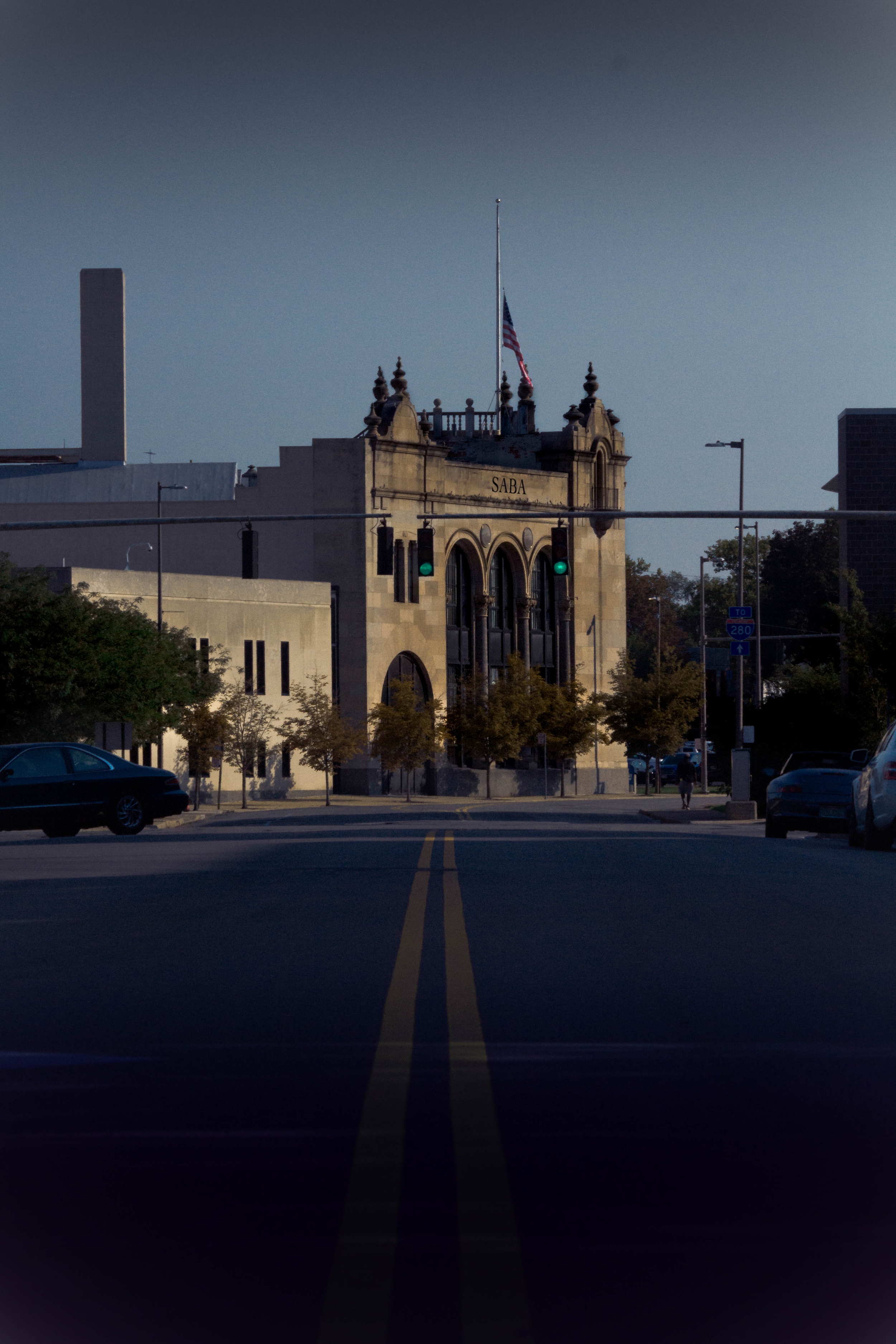 Historic building with the word 'SABA' on its facade, American flag on top, traffic lights visible, and trees in front, situated at an intersection in what appears to be downtown.