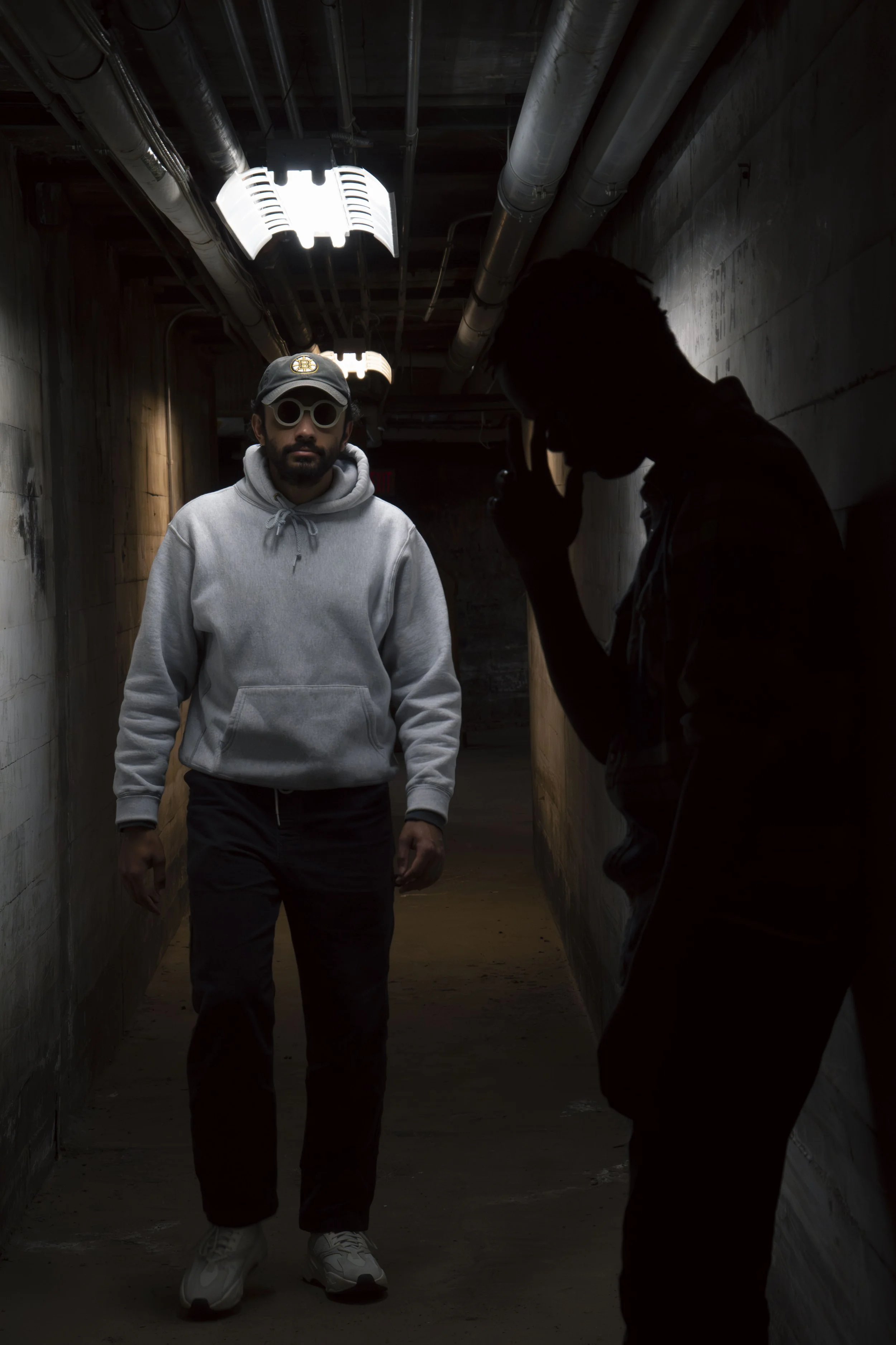A man in a gray hoodie, black pants, white sneakers, and sunglasses walking in a dimly lit industrial corridor with exposed pipes and concrete walls, with another person taking a photo in silhouette.