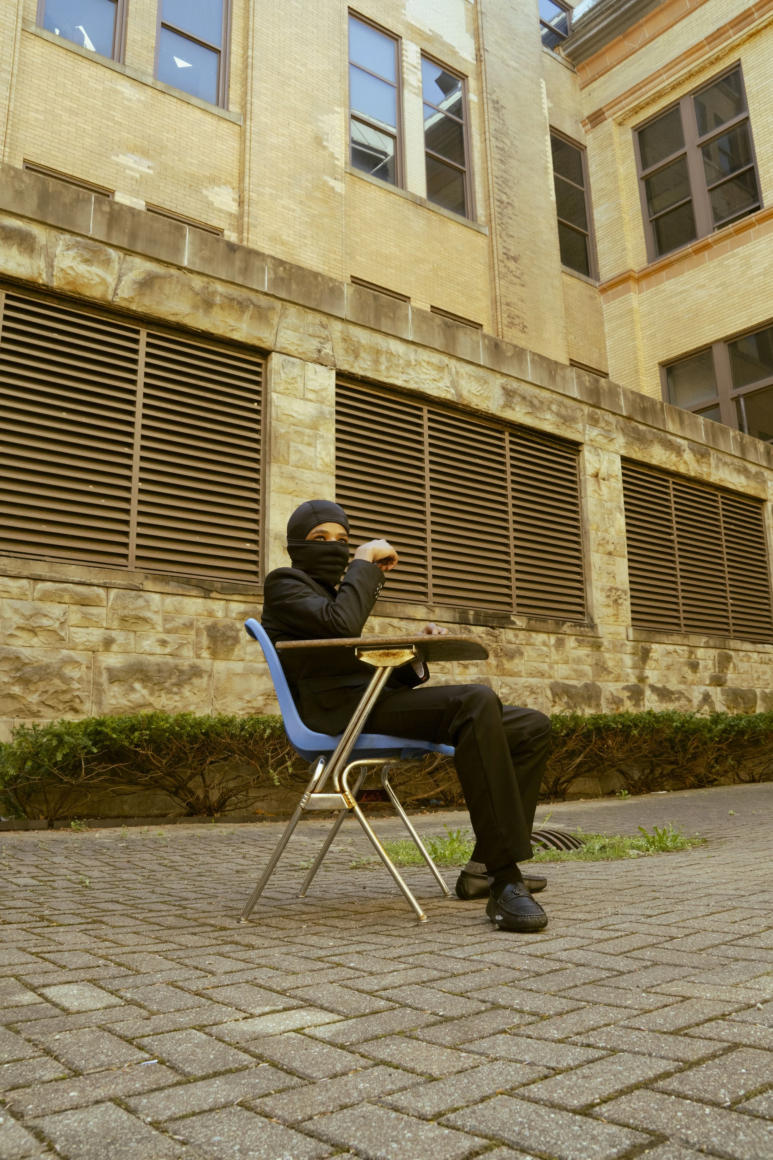 A person dressed in black clothing and a mask sitting at a school desk outside, with a backdrop of a brick building and sidewalk.