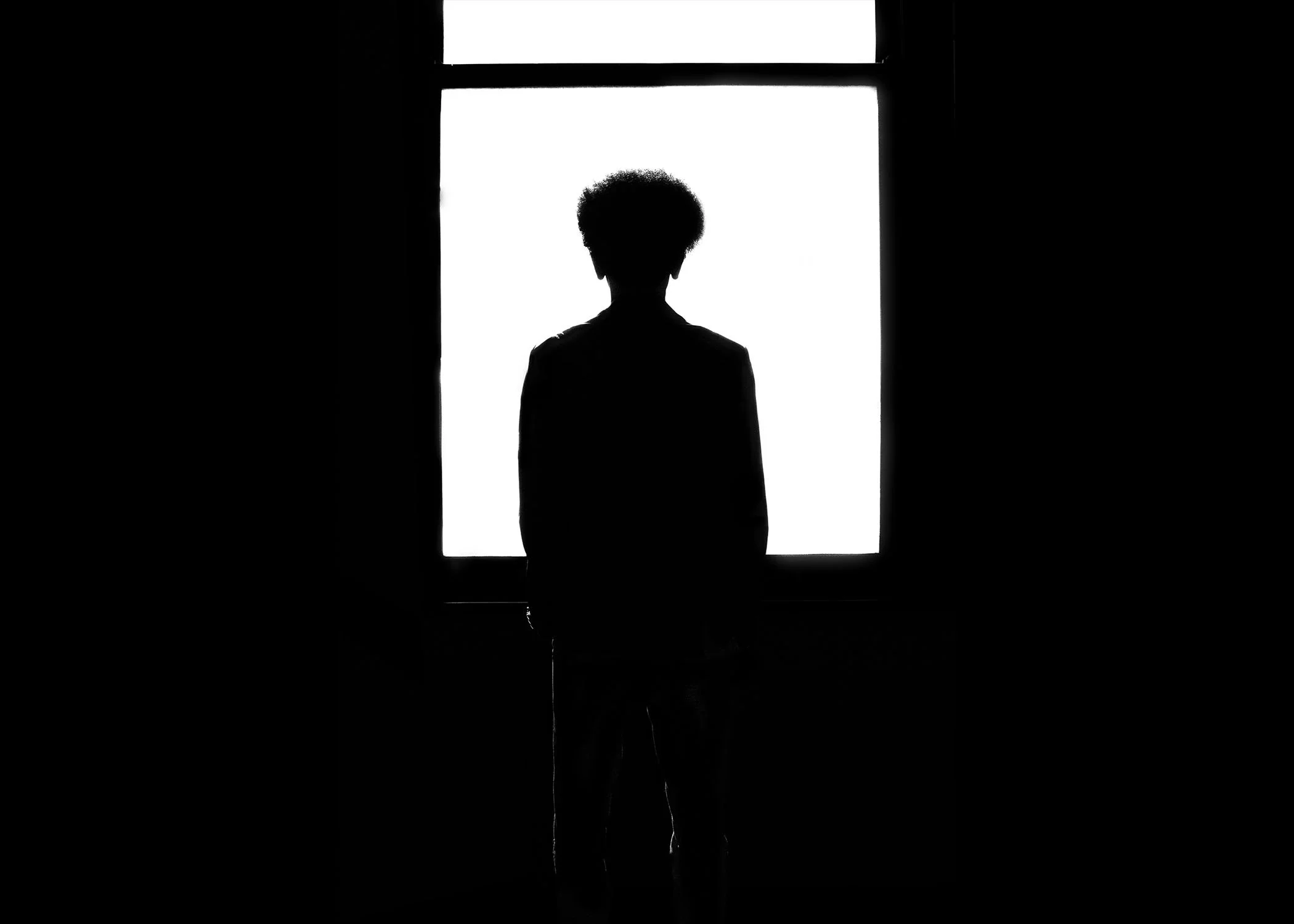 Silhouette of a person with curly hair standing by a brightly lit window, viewed from behind.