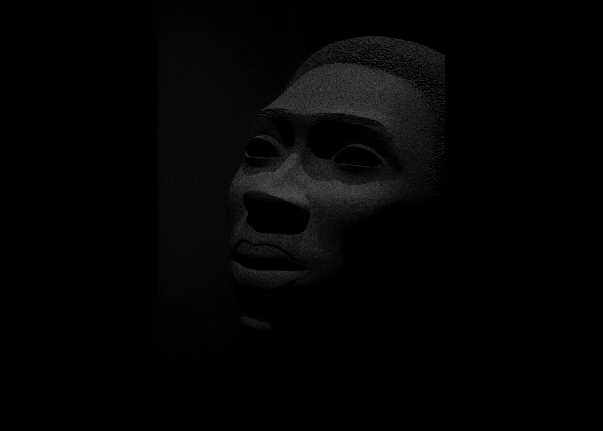 A monochrome sculpture of a human face with closed eyes, detailed eyebrows, and textured hair, set against a dark background.