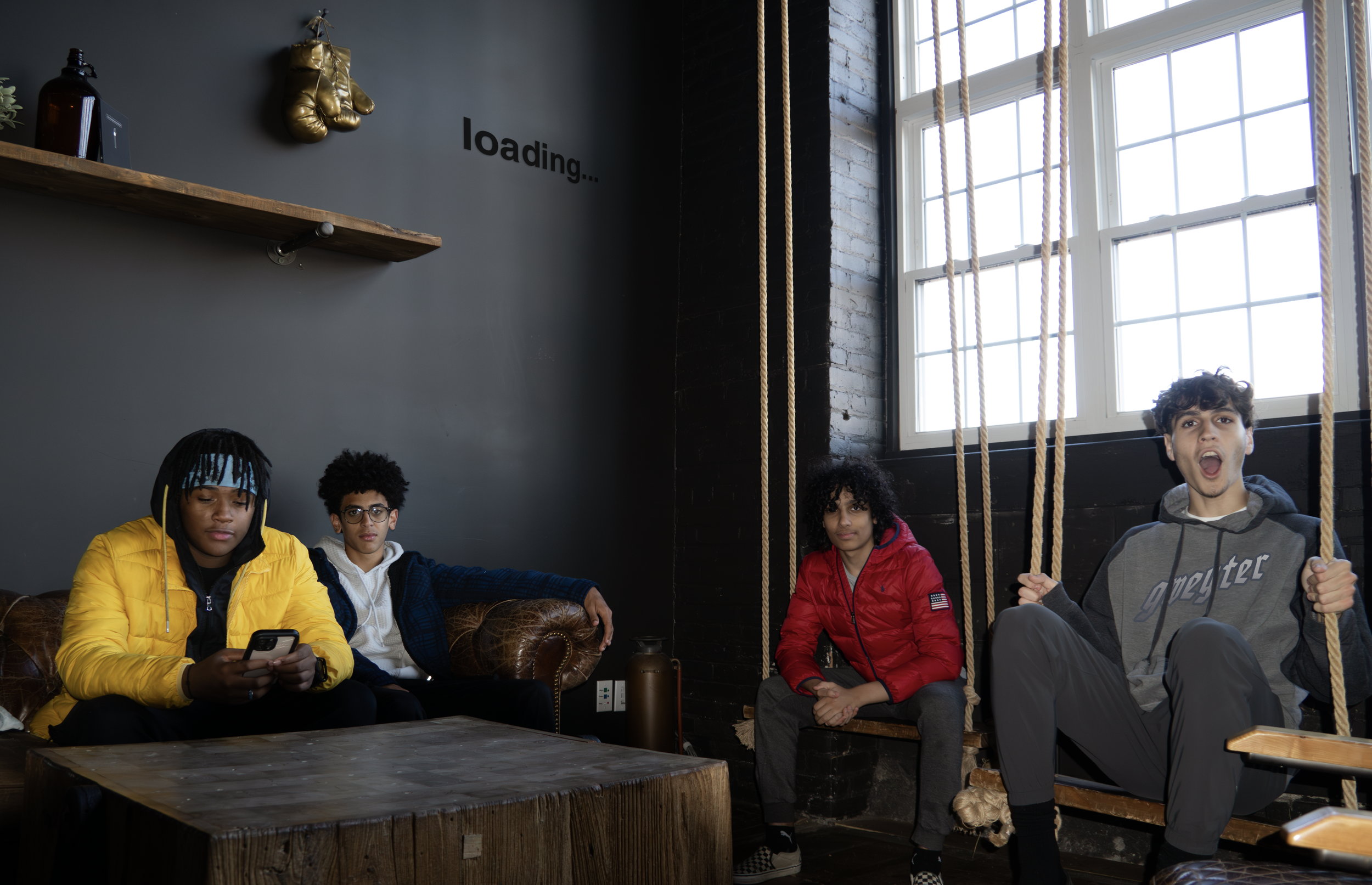 Four young people sit in a cozy, industrial-style room with large windows, a dark wall, a wooden table, and a swing. One person in a yellow jacket is looking at their phone, while the others look in various directions, with one person on the swing sh