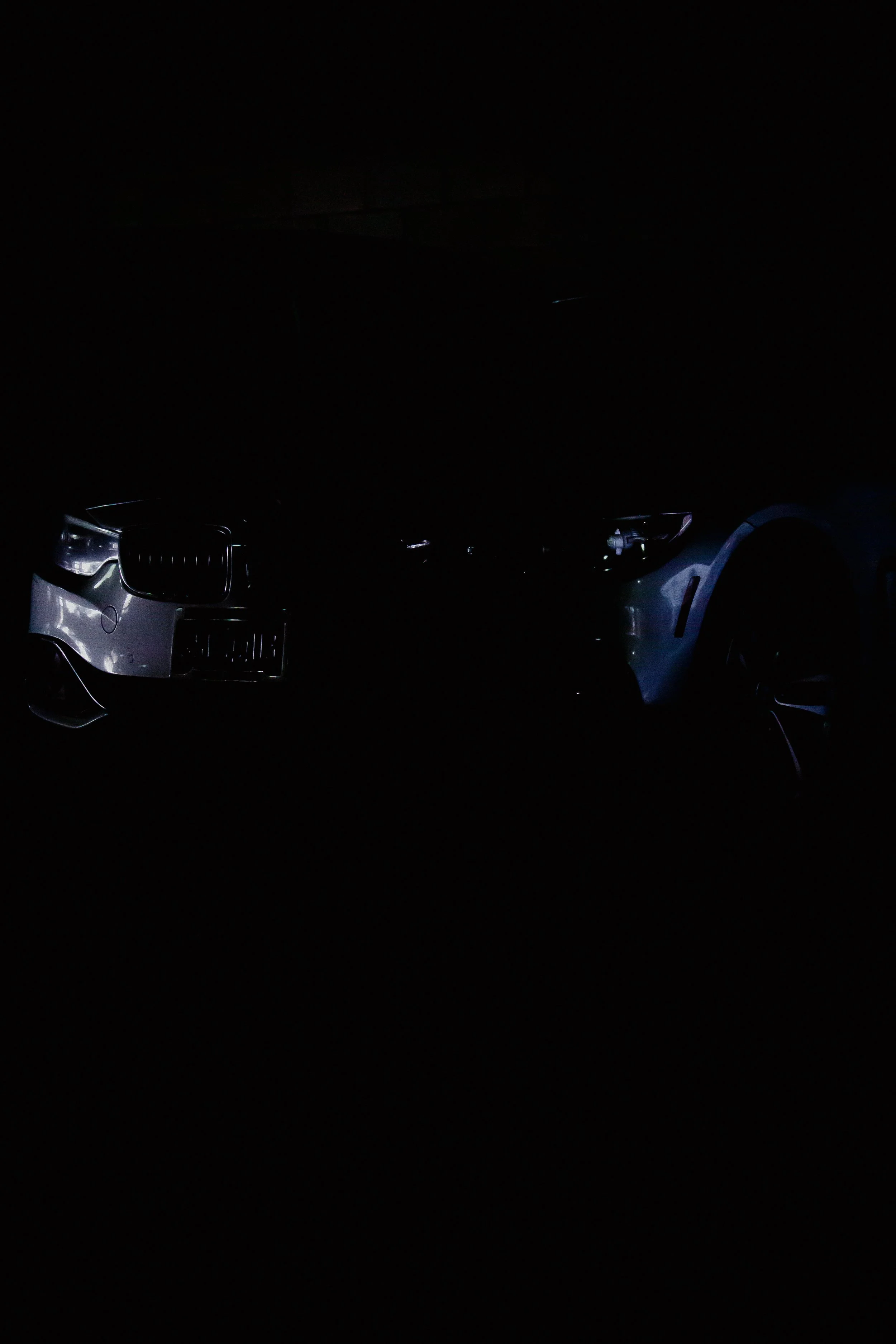 Dark image of two cars, with one on the left showing some front details and headlight, and another on the right showing a side profile, both partially illuminated.