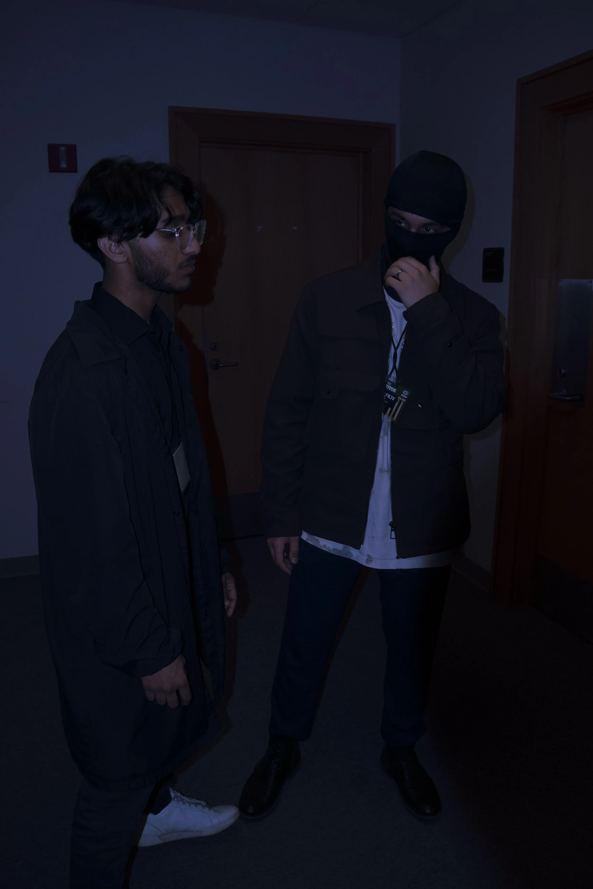 Two young men in dark clothing, one wearing glasses and the other wearing a black mask, standing in a dimly lit room near a door and a vending machine.