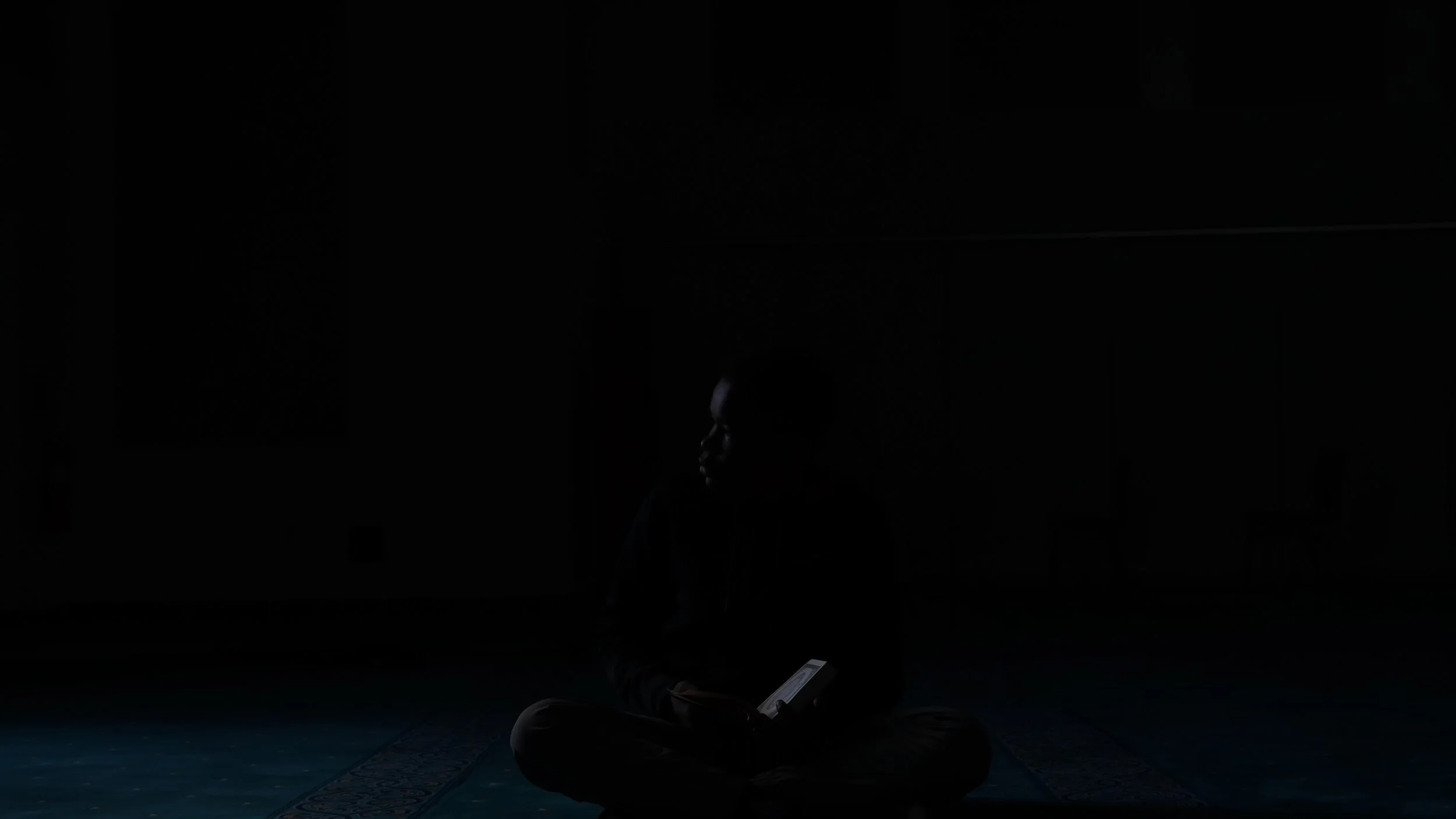 A person sitting cross-legged on the floor, illuminated by the glow of a smartphone, in a dimly lit room.