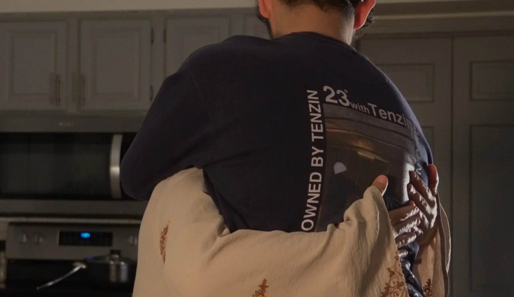 A person wearing a black T-shirt being hugged by another person wearing a cream-colored sweater in a kitchen.
