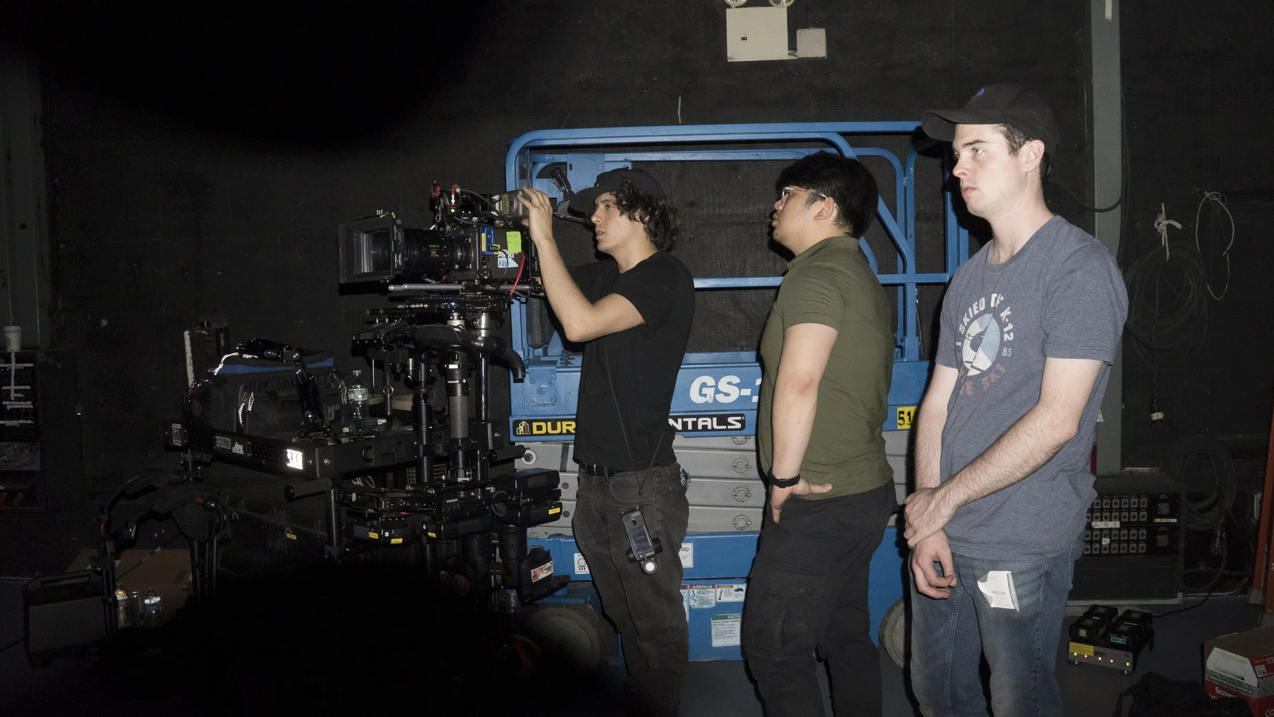 Three men on a film set stand in front of a professional camera mounted on a rig. The man on the left operates the camera, the middle man watches, and the man on the right looks forward with his arms crossed. The background includes a blue lift and v
