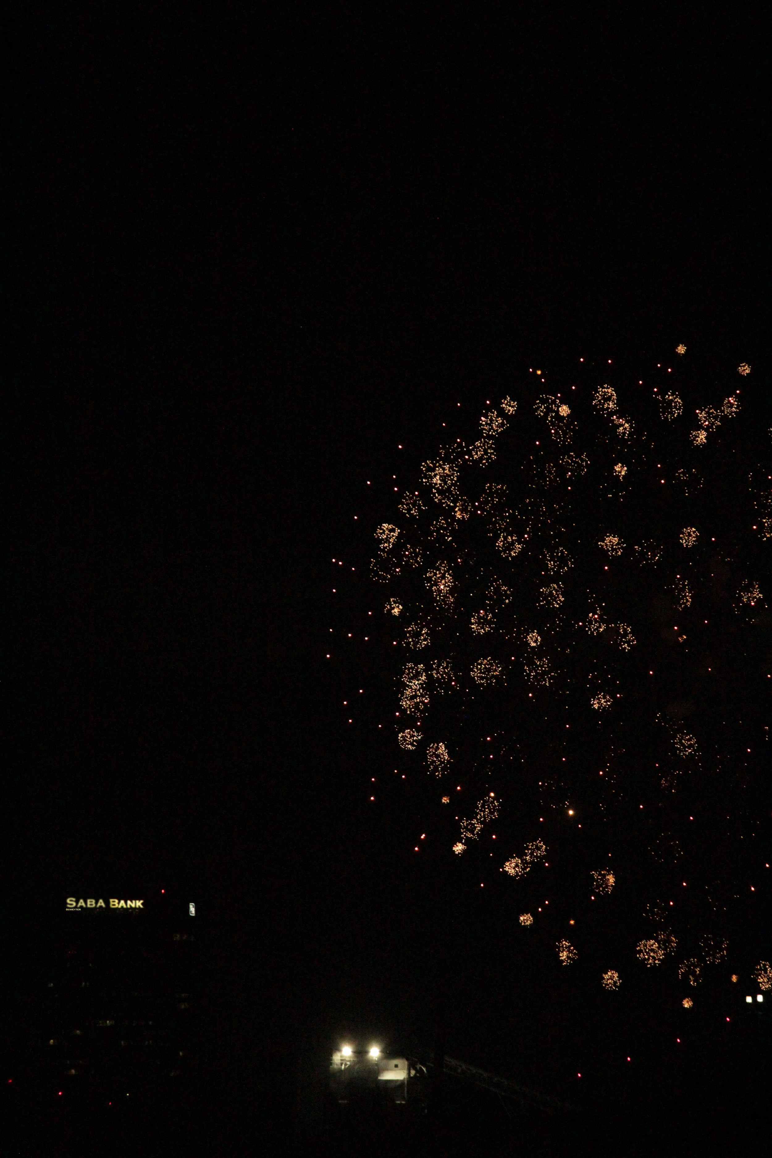 Fireworks display illuminating the night sky above a cityscape, with a building labeled 'SABA BANK' visible in the lower left corner.