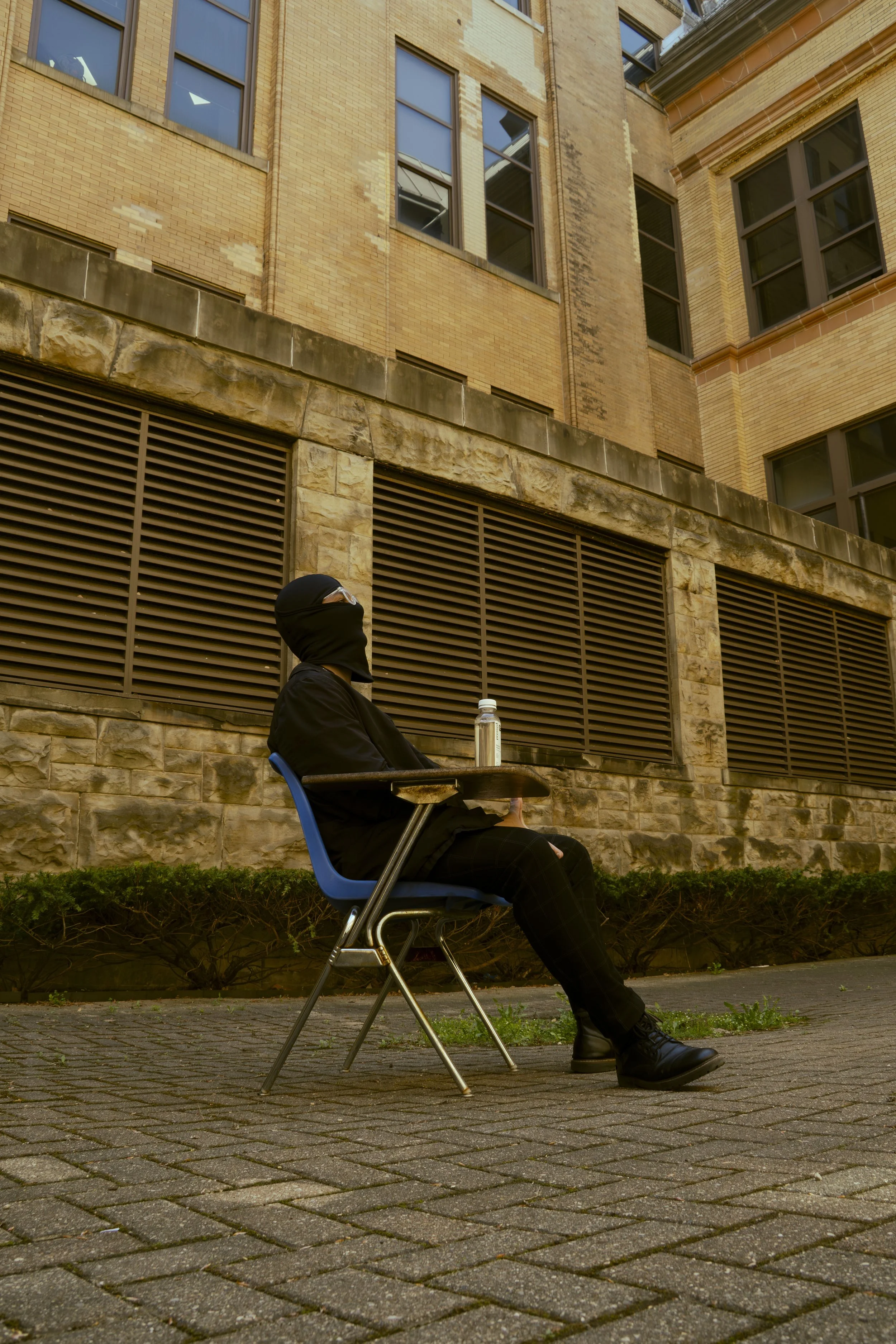 Person wearing black clothing and a ski mask sitting on a blue chair outside a brick building with large windows, with a water bottle on the table.