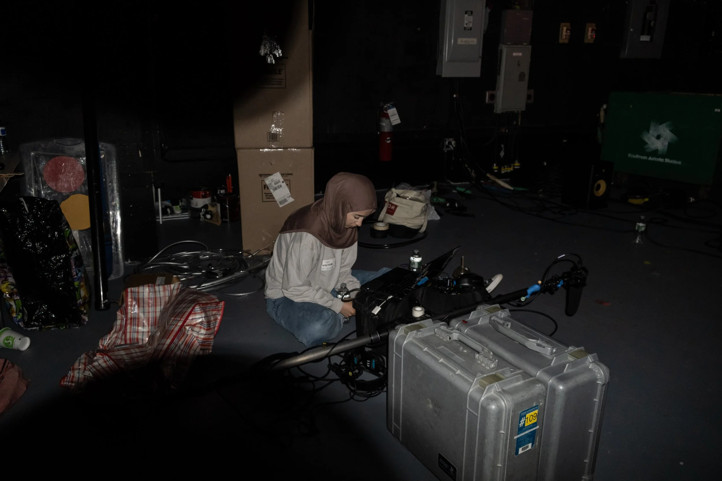 A woman sitting on a cushion on the floor surrounded by electronic equipment and cables in a dark room.
