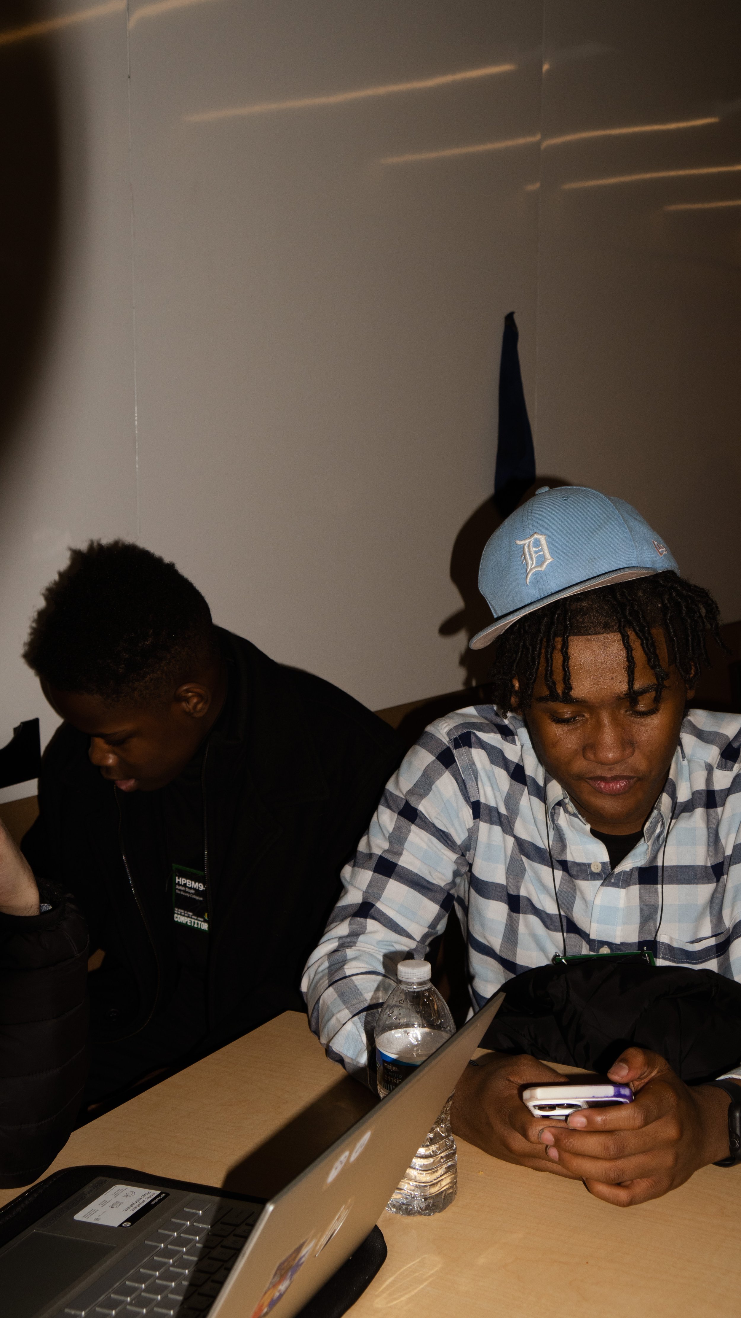 Two young men sitting at a table with laptops and water bottles, one wearing a baseball cap and the other looking down, focused on their phones.