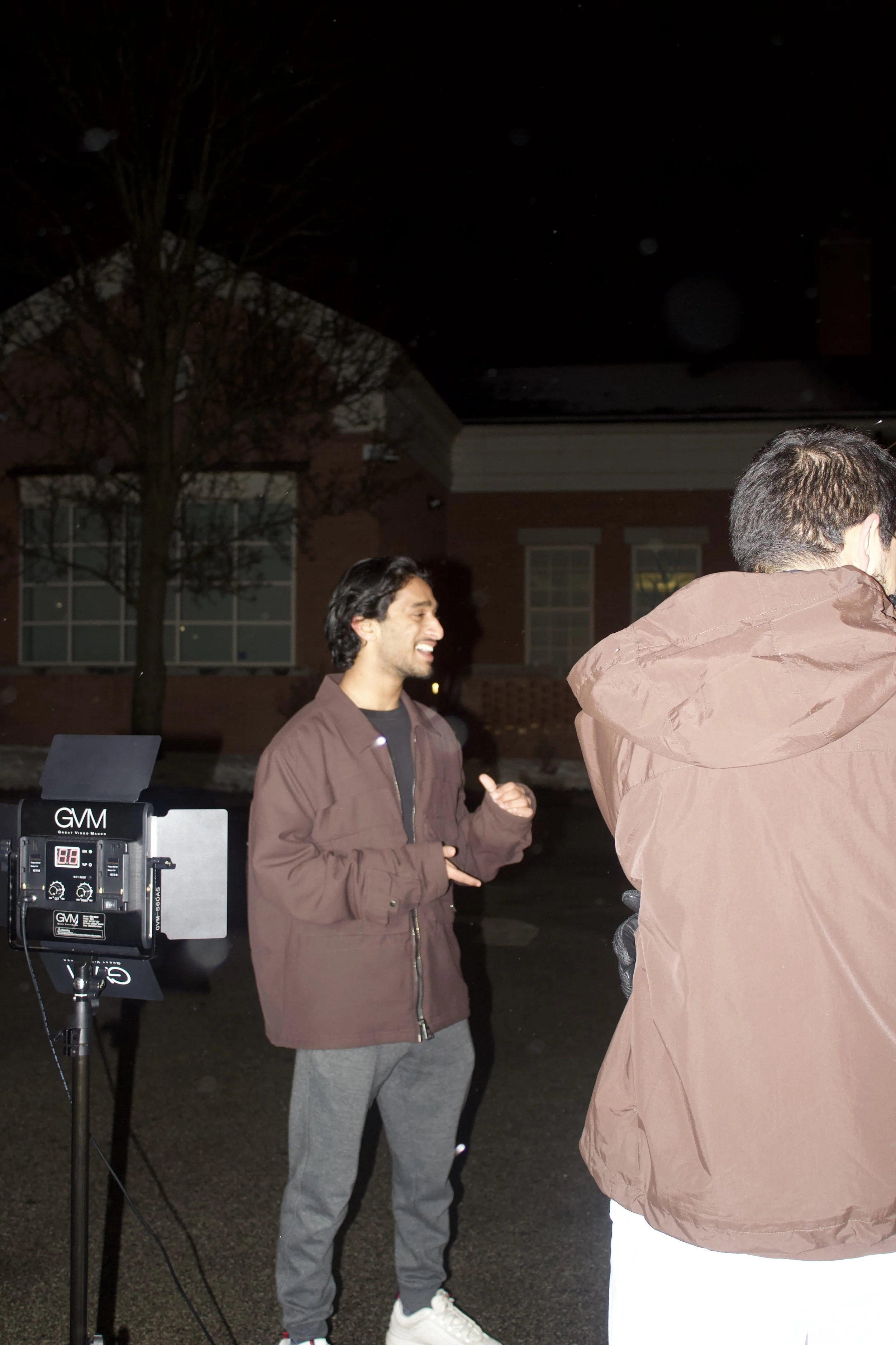 Two men standing outdoors at night, one smiling and talking, with a GVM light on a stand nearby, in front of a brick building and a tree.