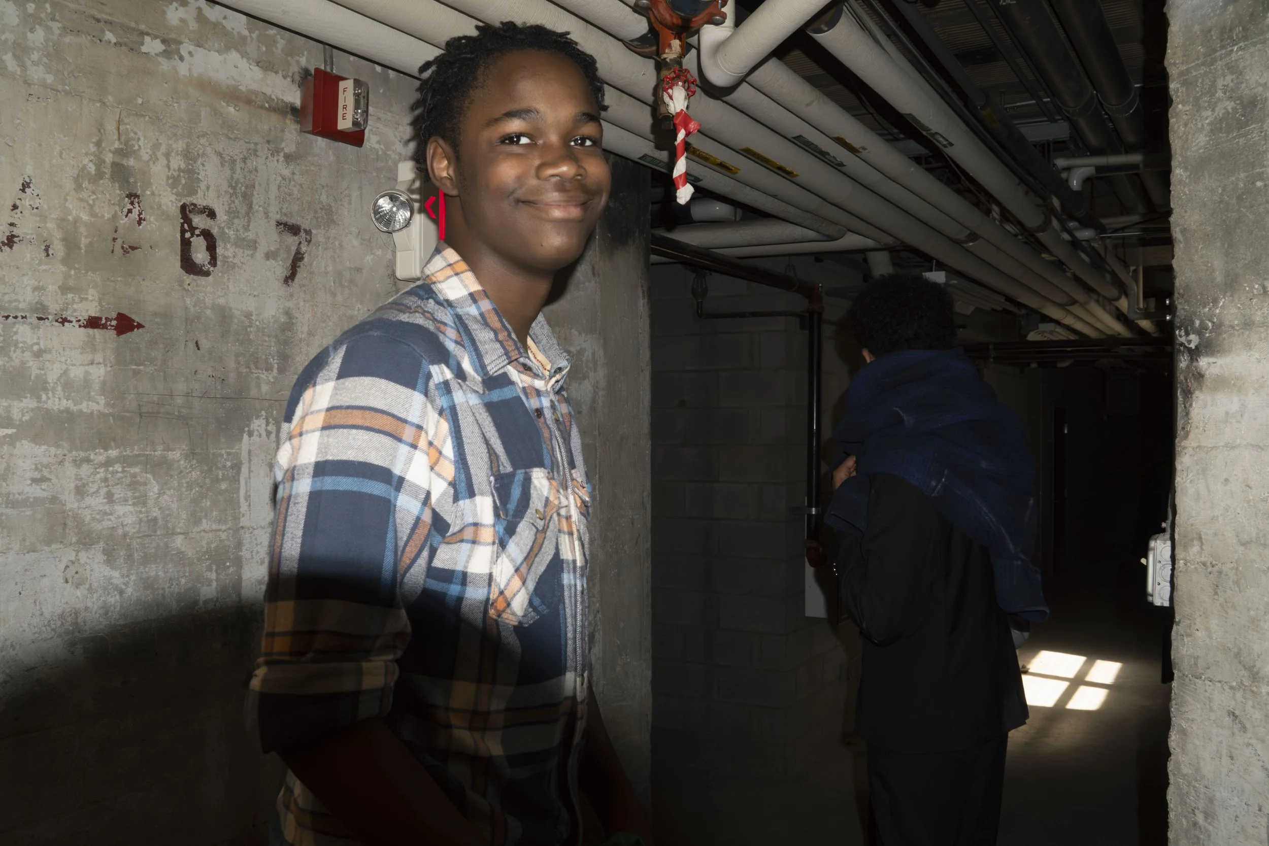 Smiling young man in a plaid shirt standing in an industrial hallway with exposed pipes and concrete walls.
