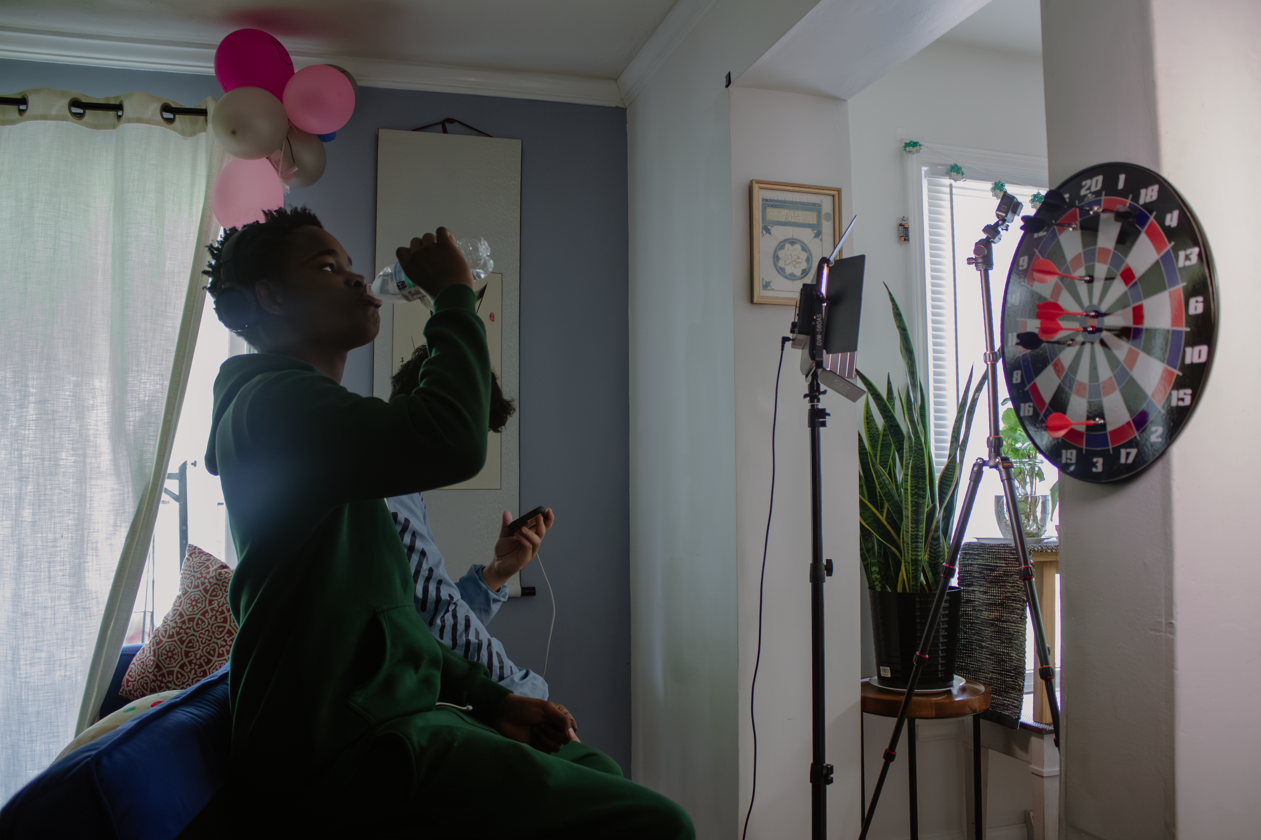 A boy in a green hoodie drinks water while sitting on a bed in a room with a dartboard and photography equipment, including a light stand and a camera, near a window with blinds and plants.