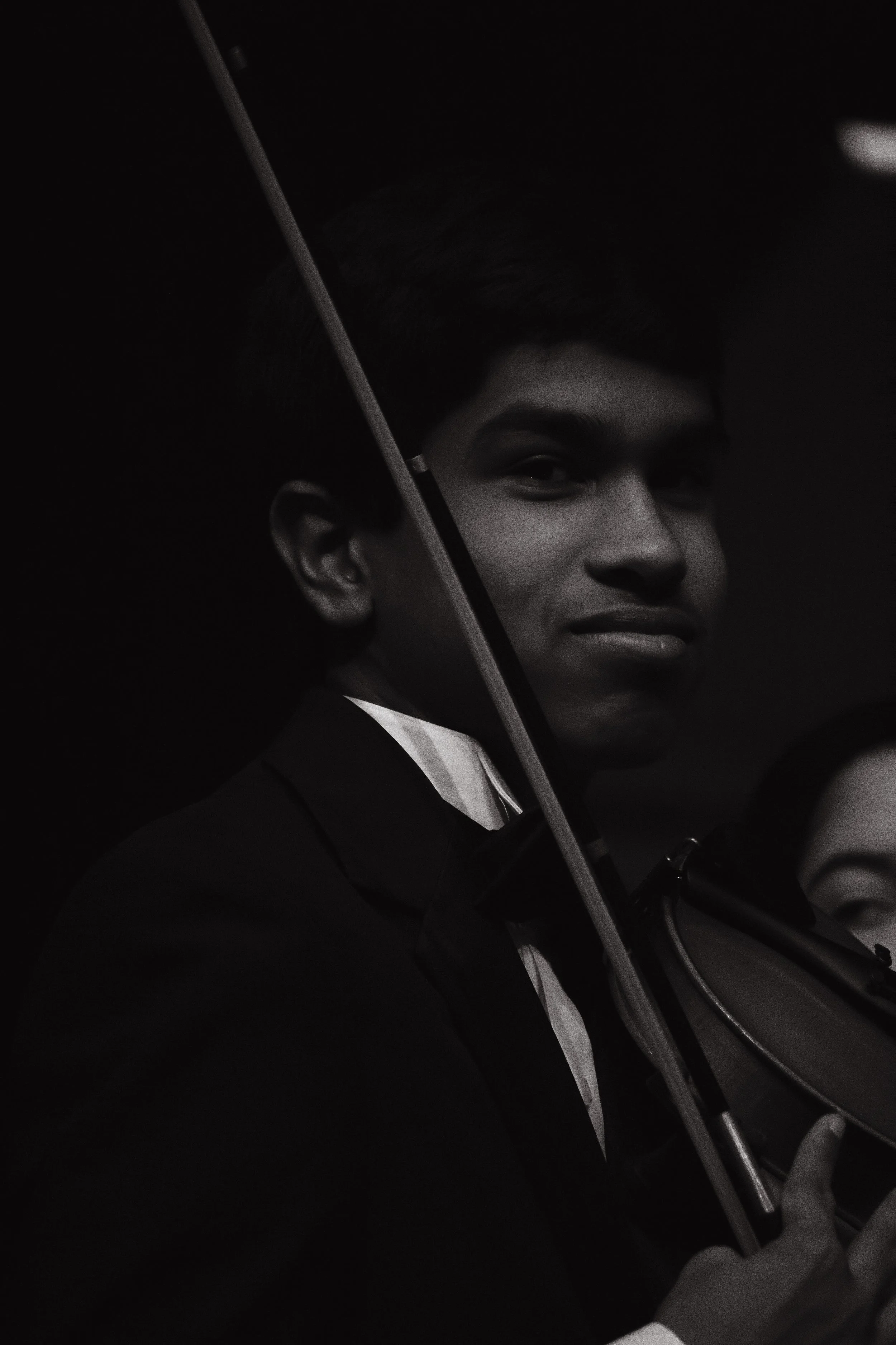 Young man holding an umbrella and a violin, dressed in formal attire, smiling at the camera in a dark environment.