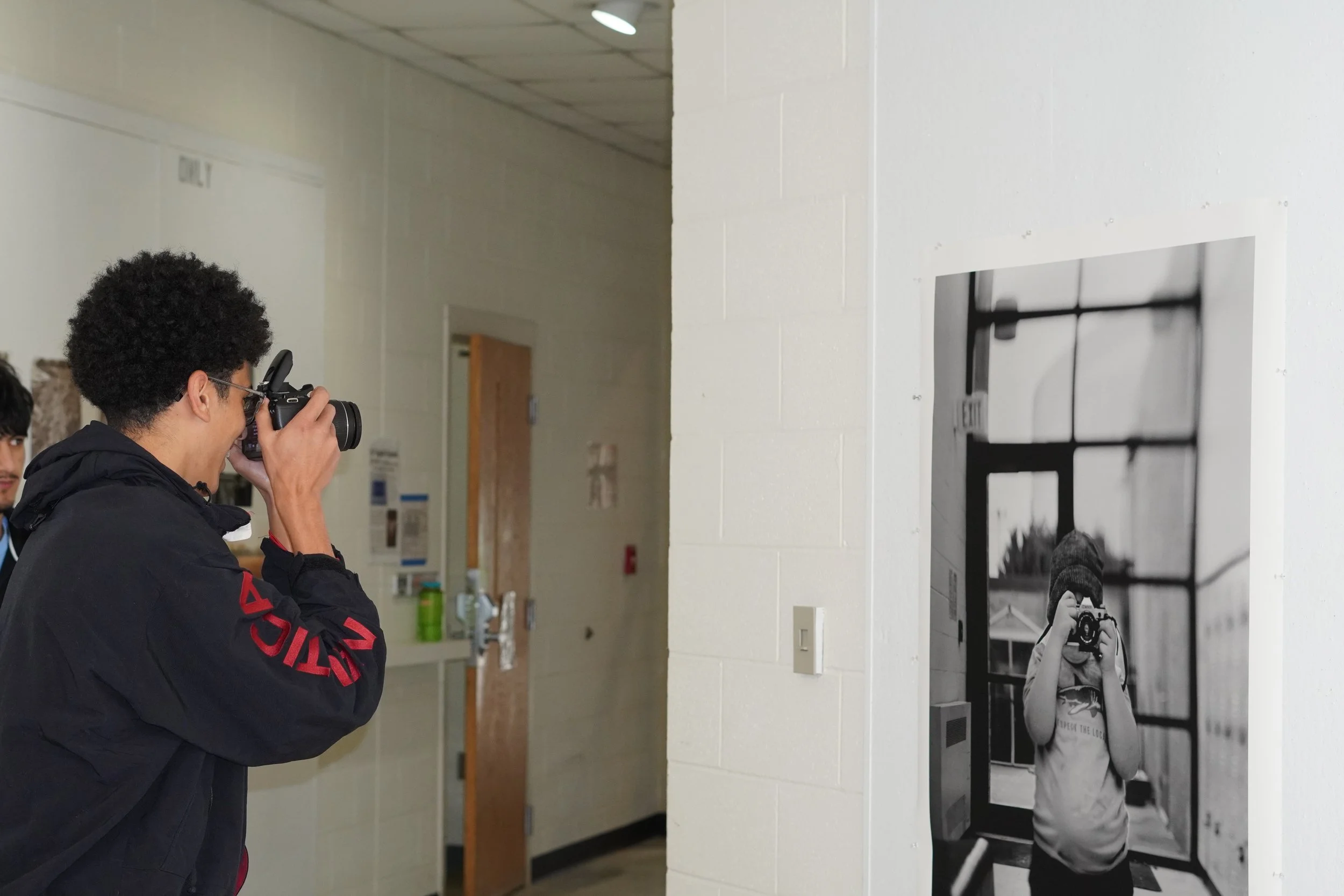 A person with curly hair wearing a black jacket is taking a photo of a black-and-white portrait on a gallery wall. The portrait shows a young person with a camera, wearing a hoodie and a beanie, standing in front of large windows.