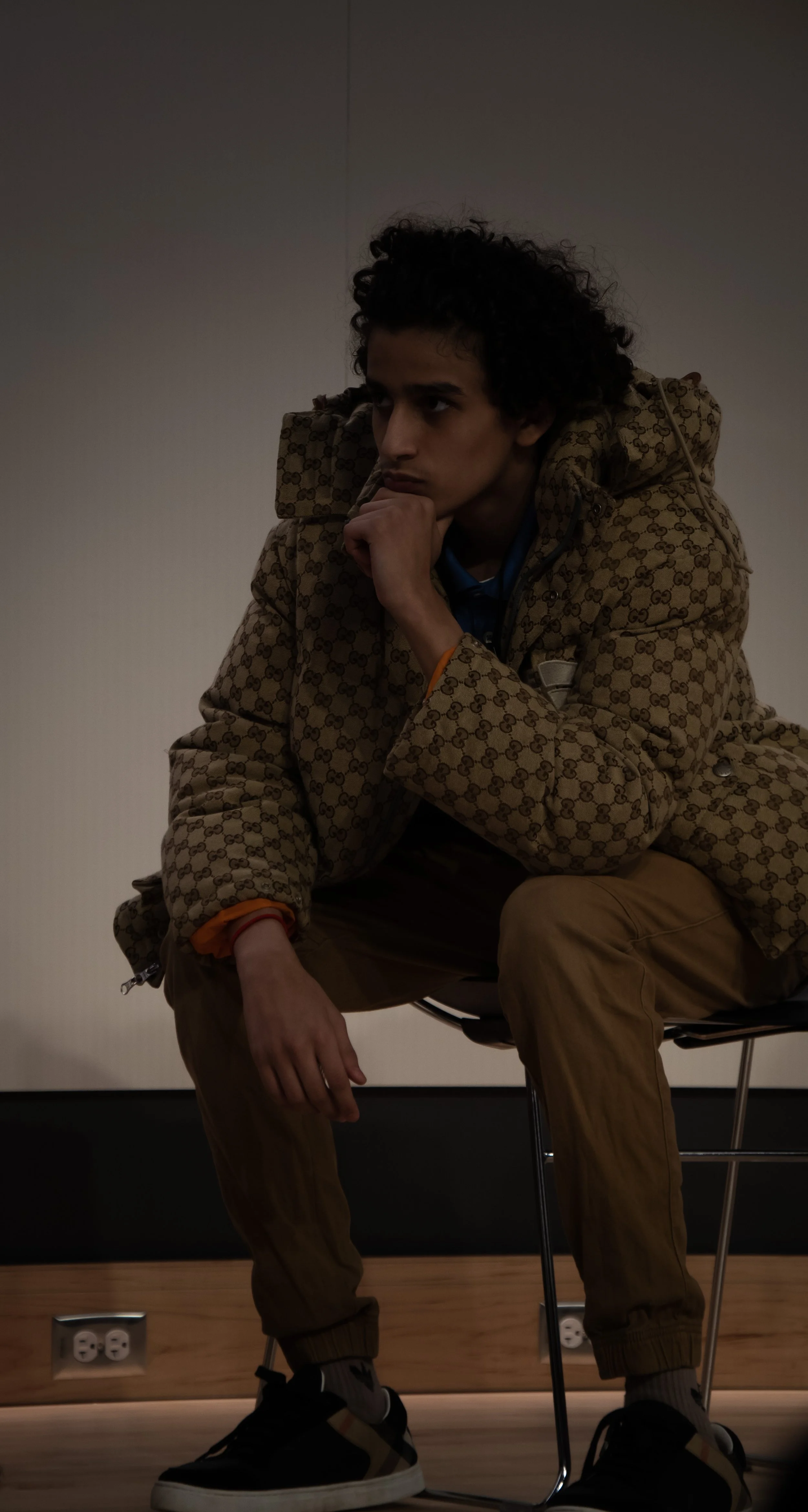 A young man with curly hair sitting on a black chair, wearing a beige patterned jacket, khaki pants, and black sneakers, in a contemplative pose with his chin resting on his hand in a room with a plain wall and two electrical outlets.