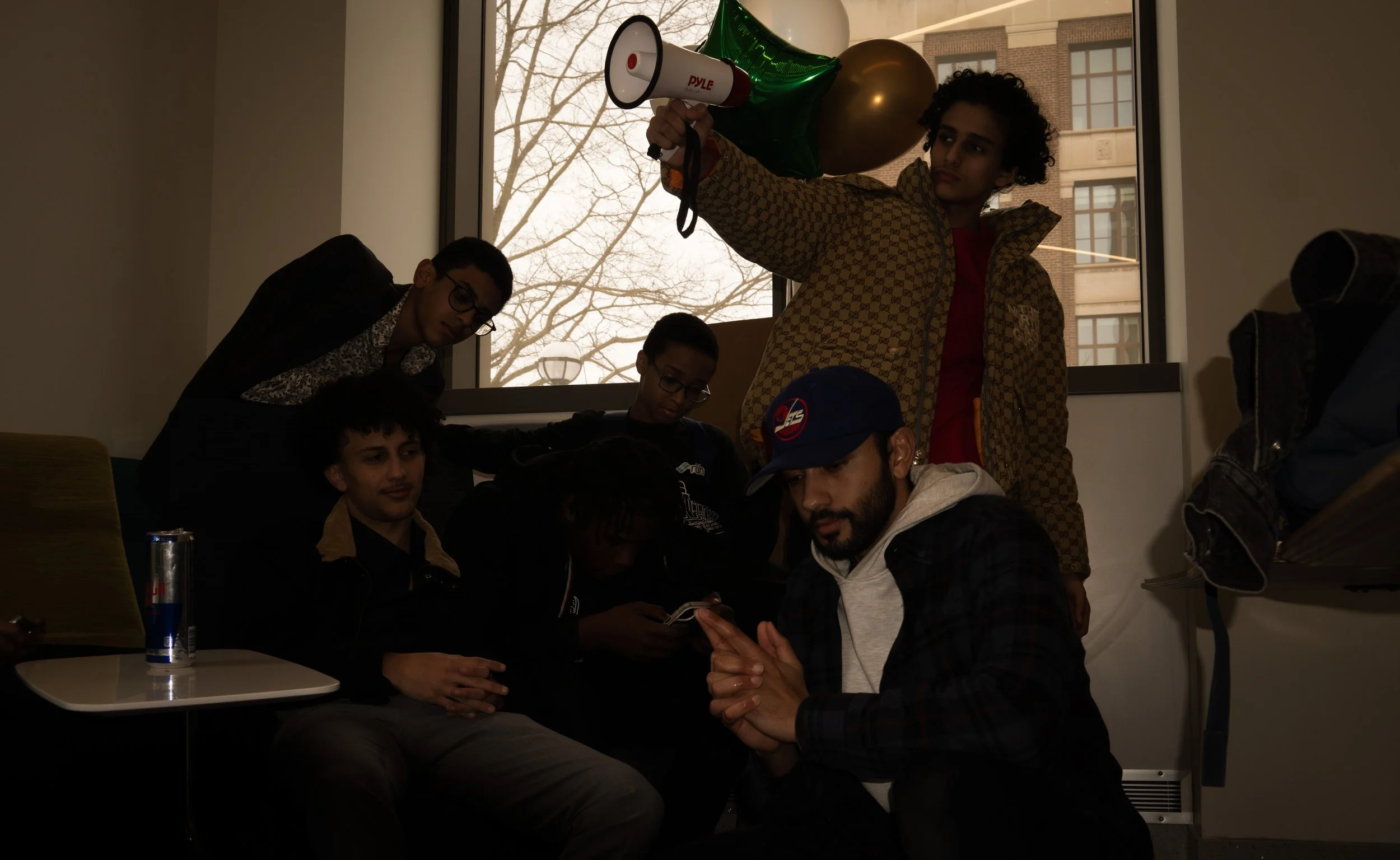 Group of young people gathered in a room near a window, some sitting and some standing, with balloons and a person holding a megaphone.