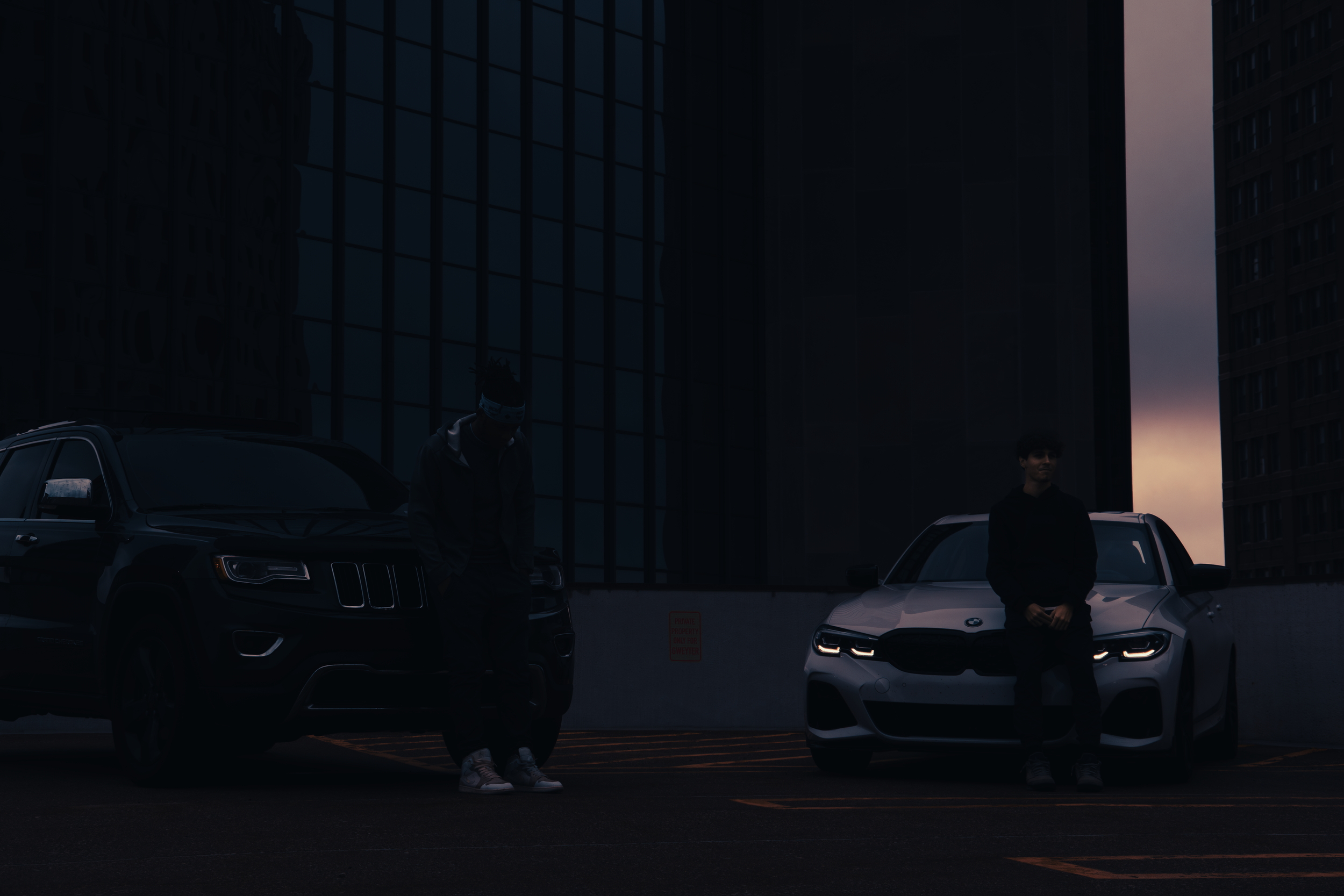 Two men standing in front of parked cars on a rooftop parking lot at dusk, with dark urban buildings in the background.