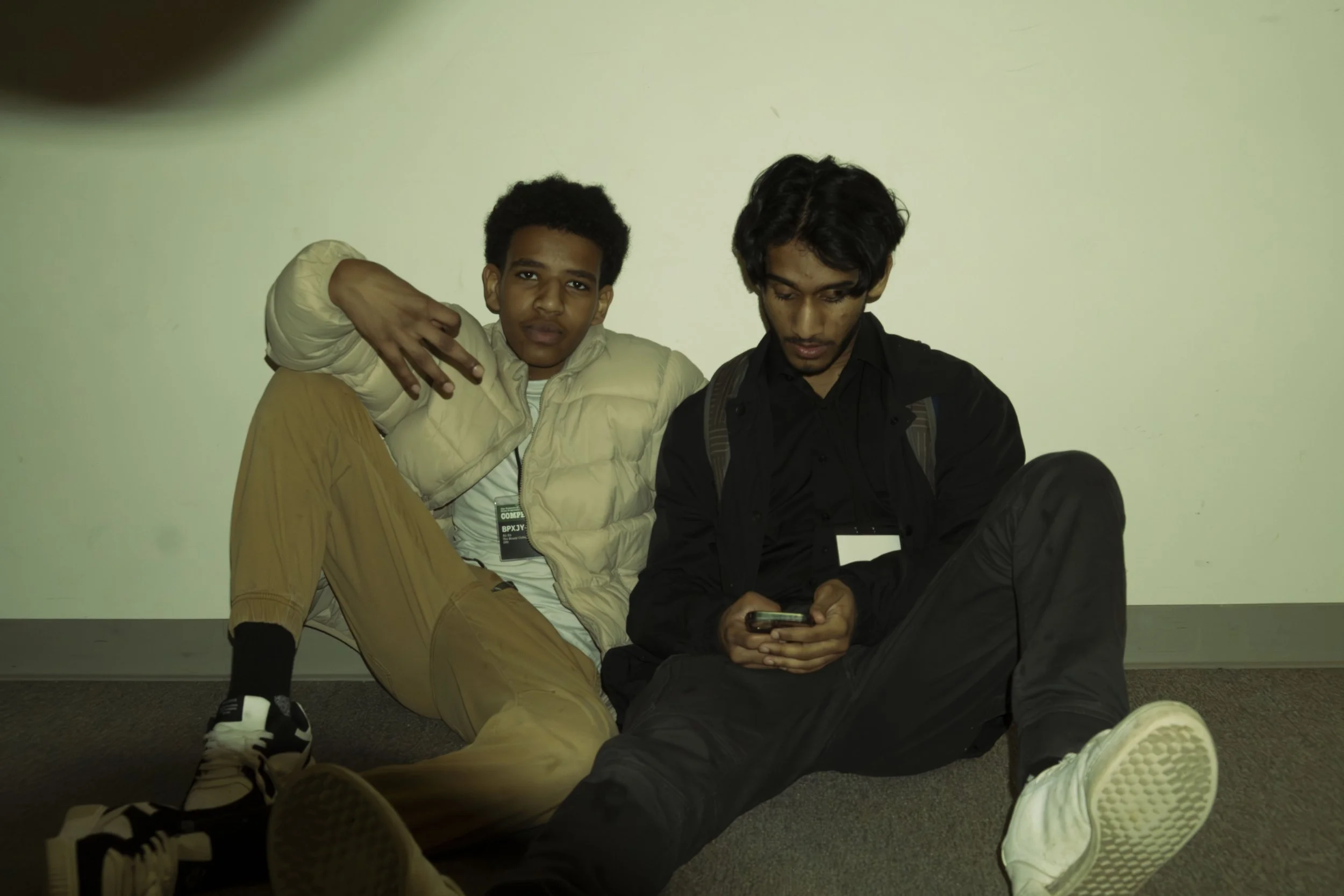 Two young men sitting on the floor against a plain wall, one wearing a beige jacket and the other in black, with one looking at a phone and the other looking at the camera.