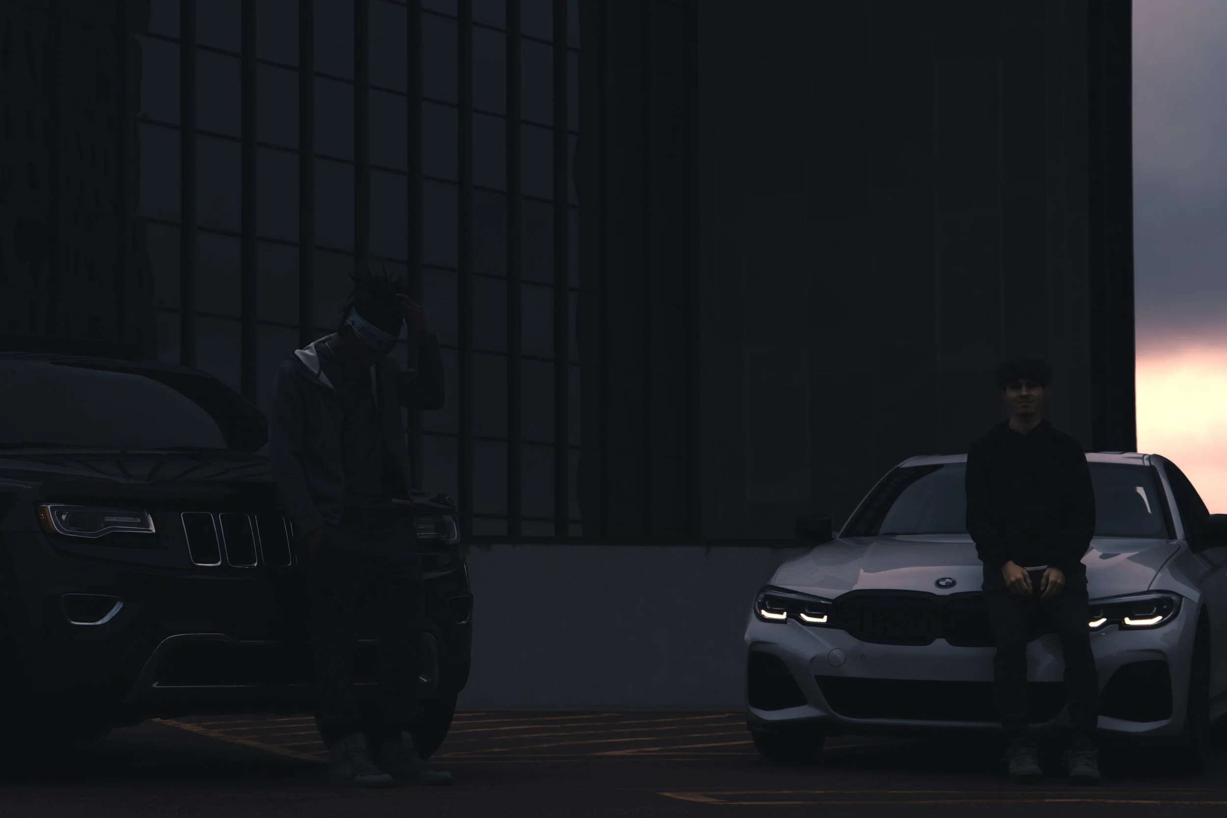 Two people standing between black and white cars in front of a modern building at sunset or dusk.