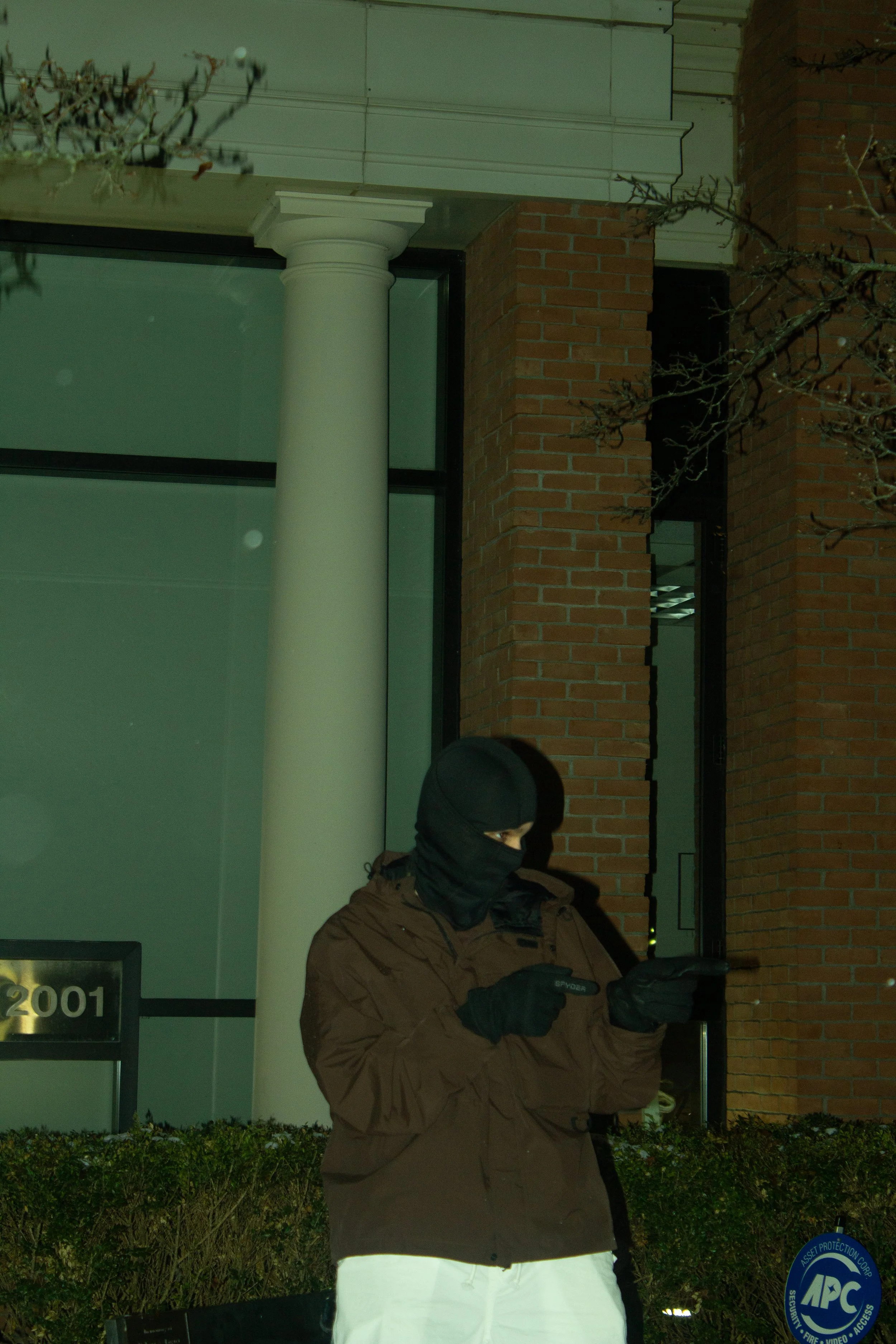 A person dressed in dark clothing, wearing a mask and gloves, using a mobile phone on a sidewalk outside a building with brick walls and large glass windows at night.