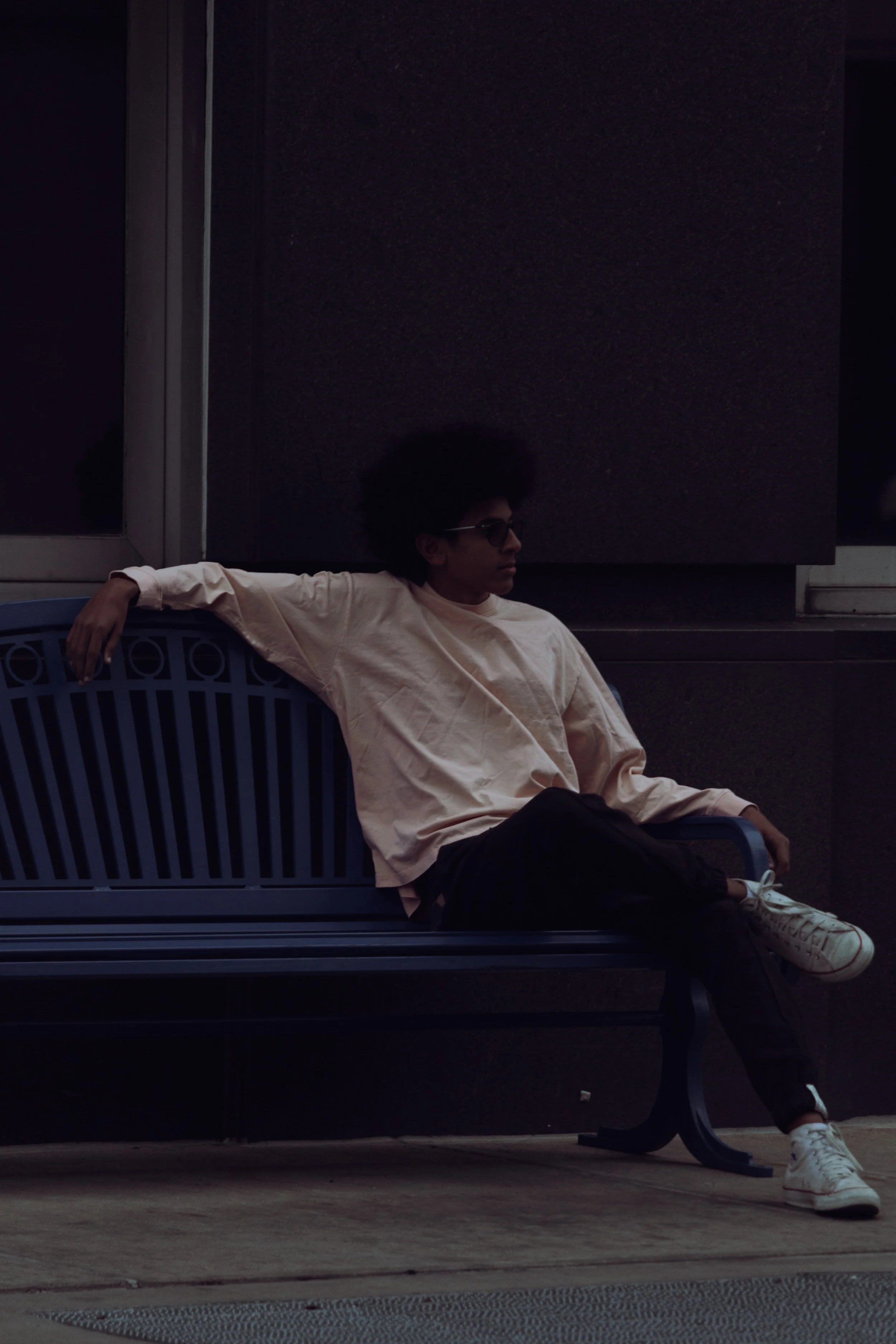 A young person with sunglasses and an afro hairstyle sitting on a dark blue bench in a dimly lit urban setting.