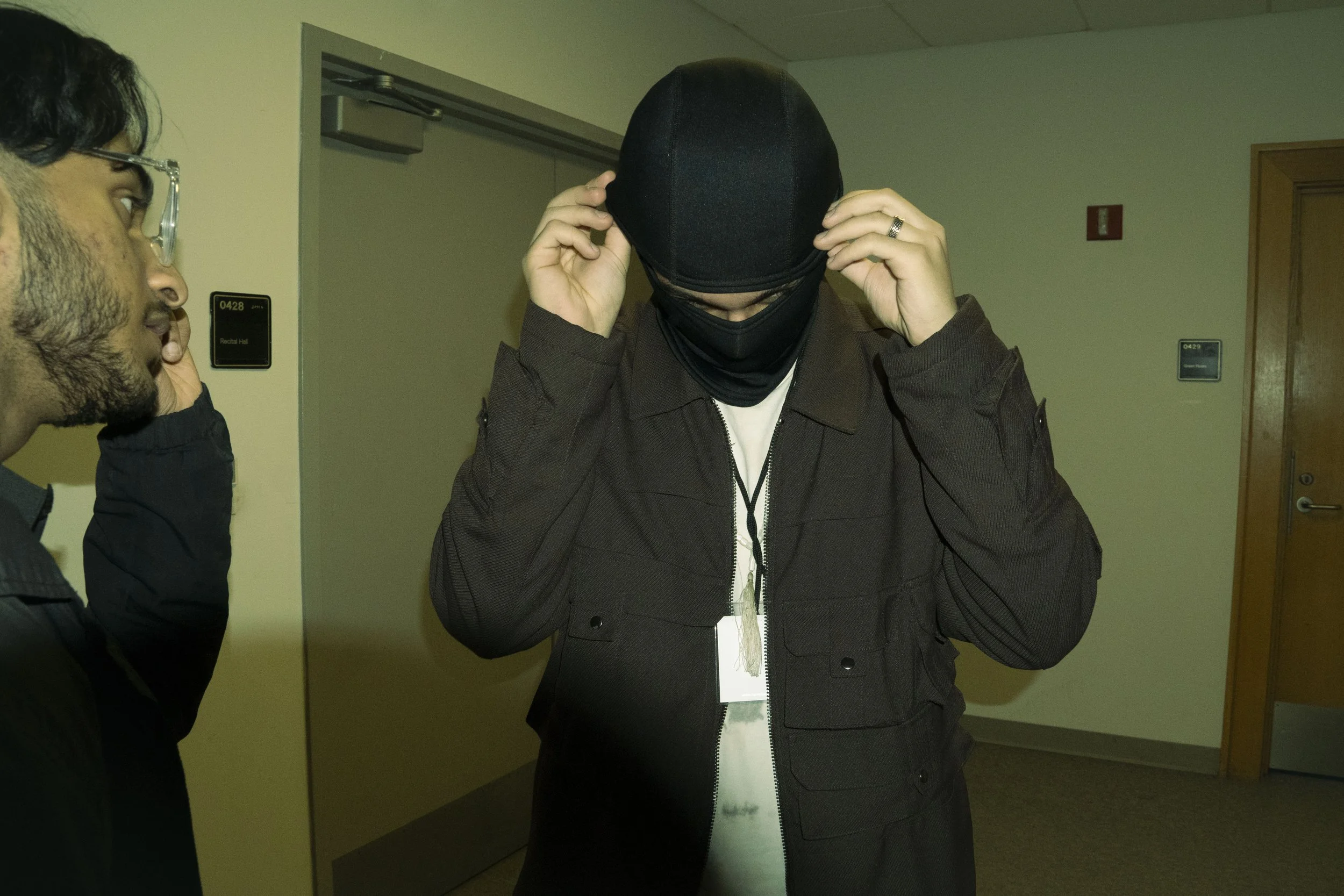 A person wearing a black ski mask and dark jacket adjusting the mask with both hands, standing indoors. Another person with glasses and facial hair is to the left, observing.