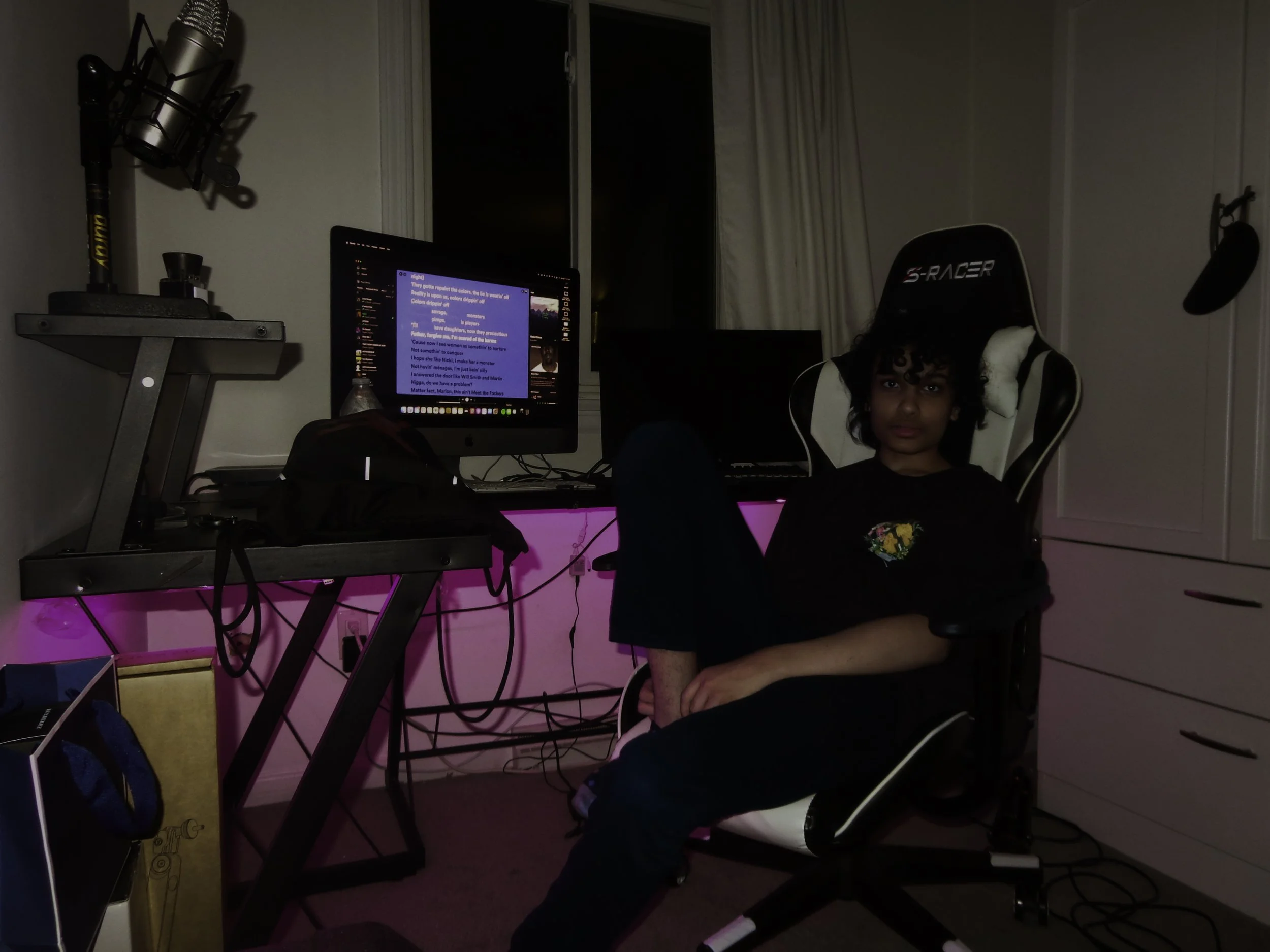 Person sitting in a gaming chair in front of a computer desk with dual monitors, one displaying a chat application, a microphone mounted on a stand, and pink LED lighting under the desk.