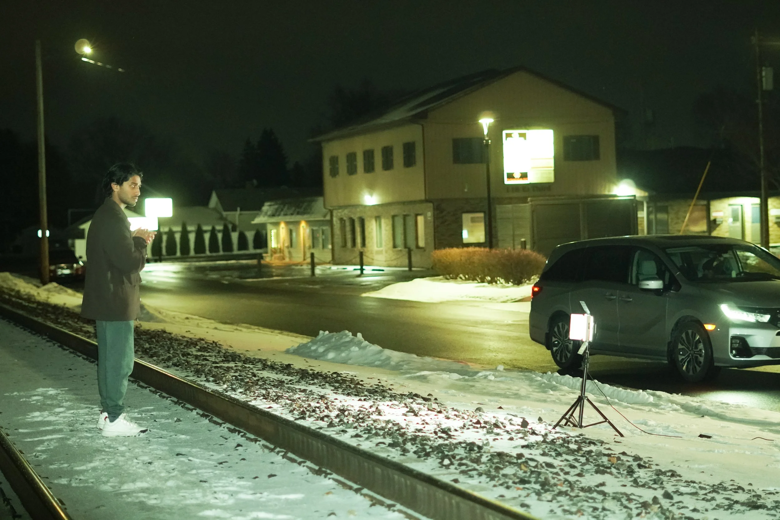 A young man in a dark jacket and light green pants standing on snow-covered ground near railroad tracks at night, looking at a bright light on a tripod, with a parked gray car and buildings illuminated in the background.