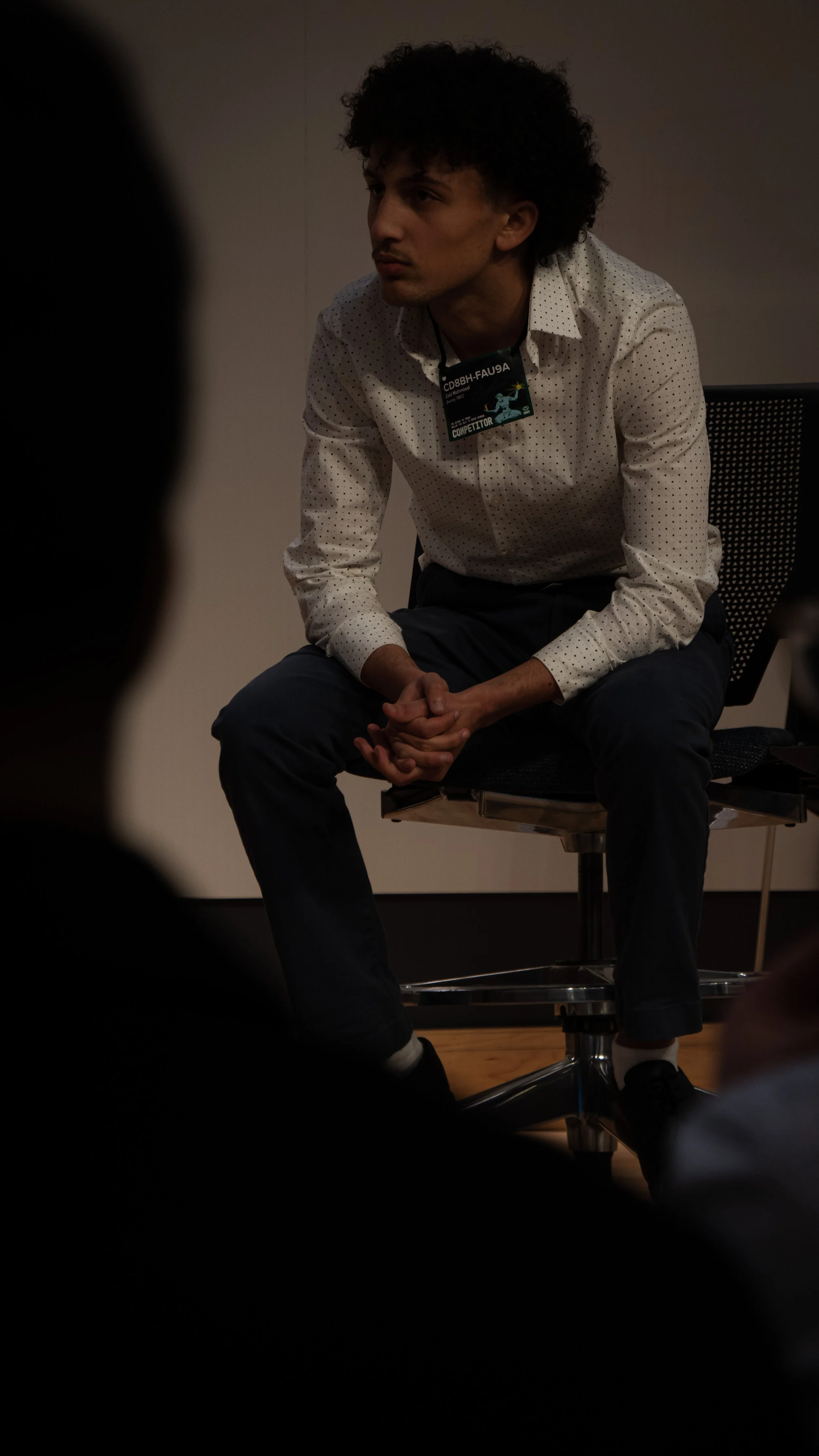 A young man with curly hair, wearing a white shirt with small black dots, is sitting on a chair during an event or discussion, with his hands clasped together.