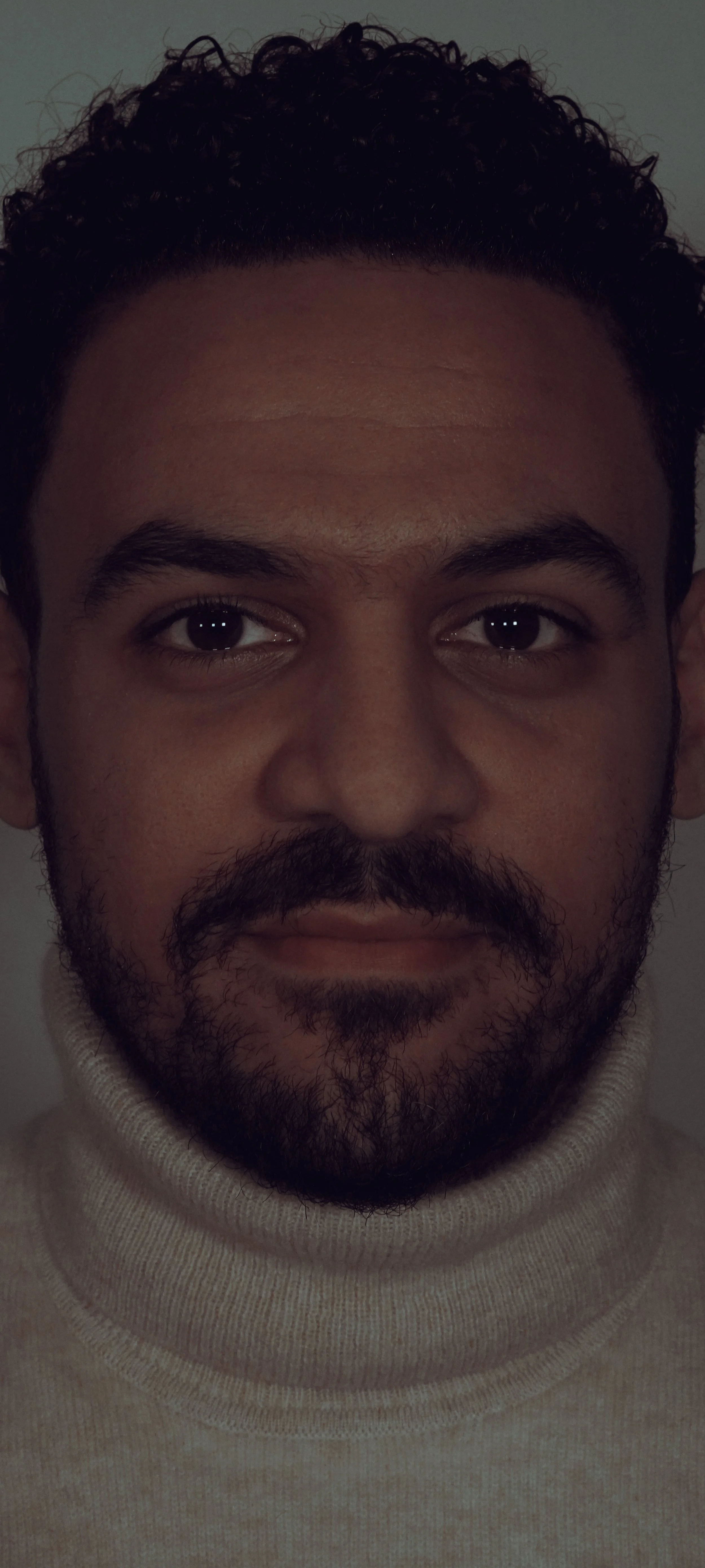 Close-up portrait of a man with dark curly hair, a beard and mustache, wearing a light-colored turtleneck sweater.