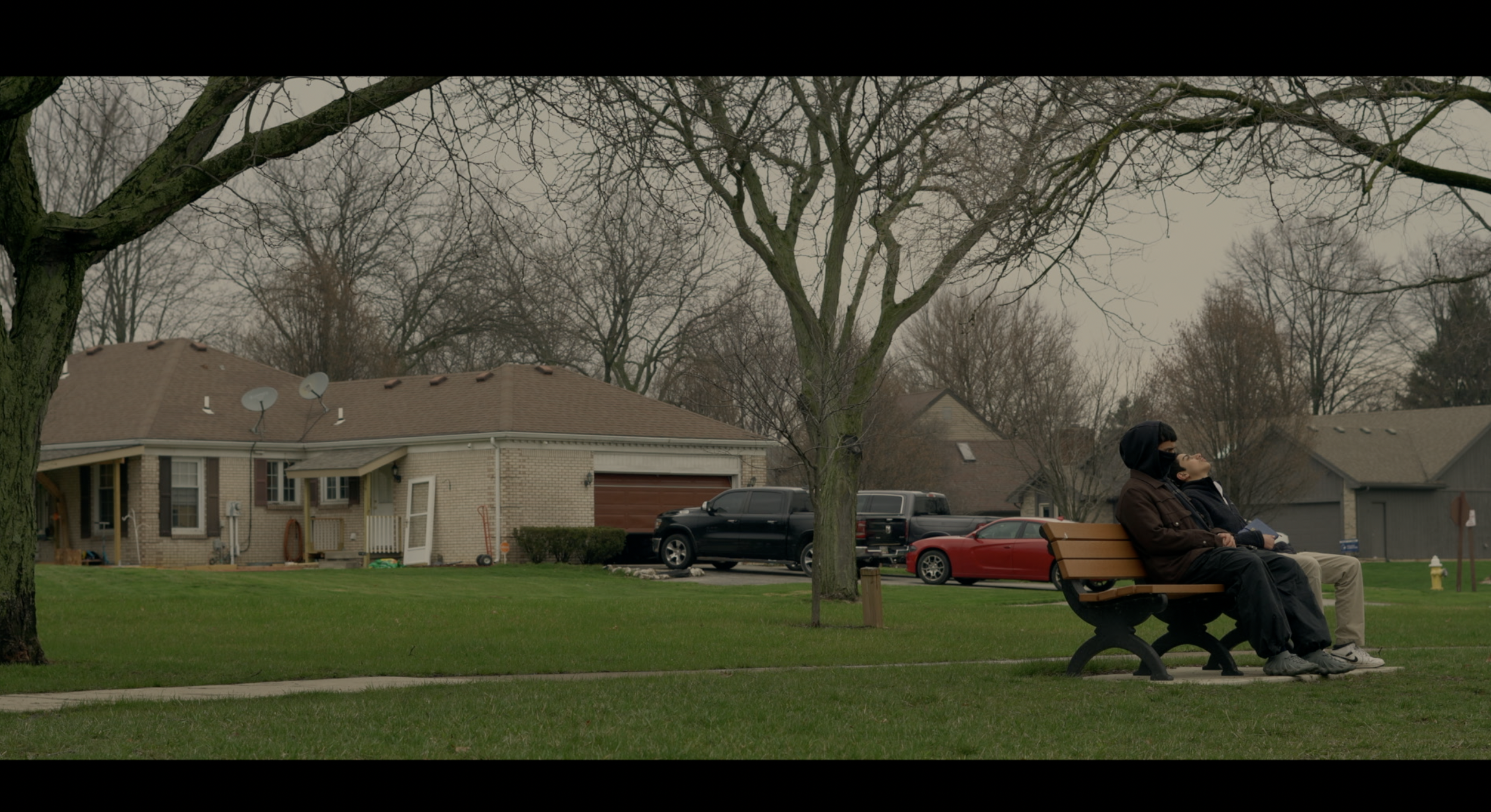 Two people sitting on a park bench under leafless trees in front of houses with cars parked in the driveway.