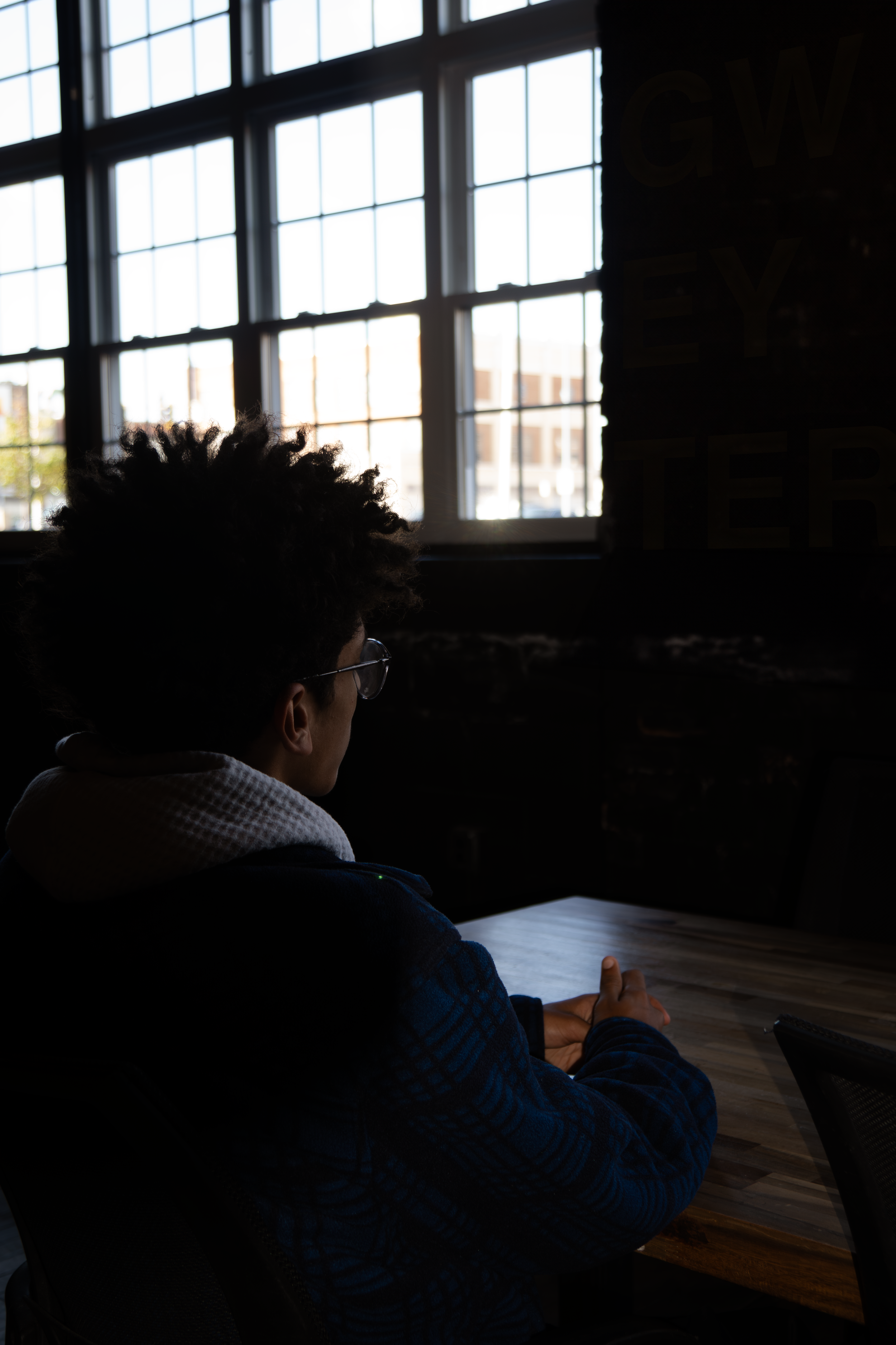 Person sitting at a table in a dark room, facing window with natural light coming in.