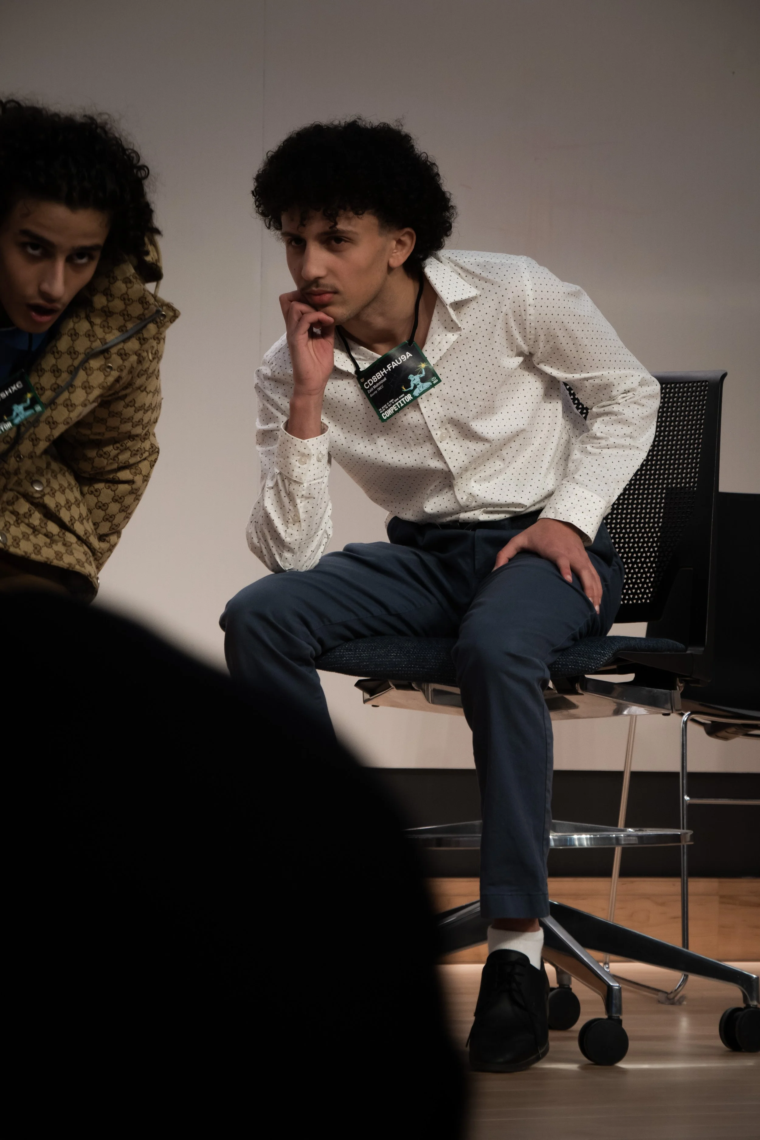 Two young men sitting on chairs, one appears to be thinking or concentrating while the other leans in to look at something off-camera. They are in a room with plain walls, and each wears a conference badge around their neck.