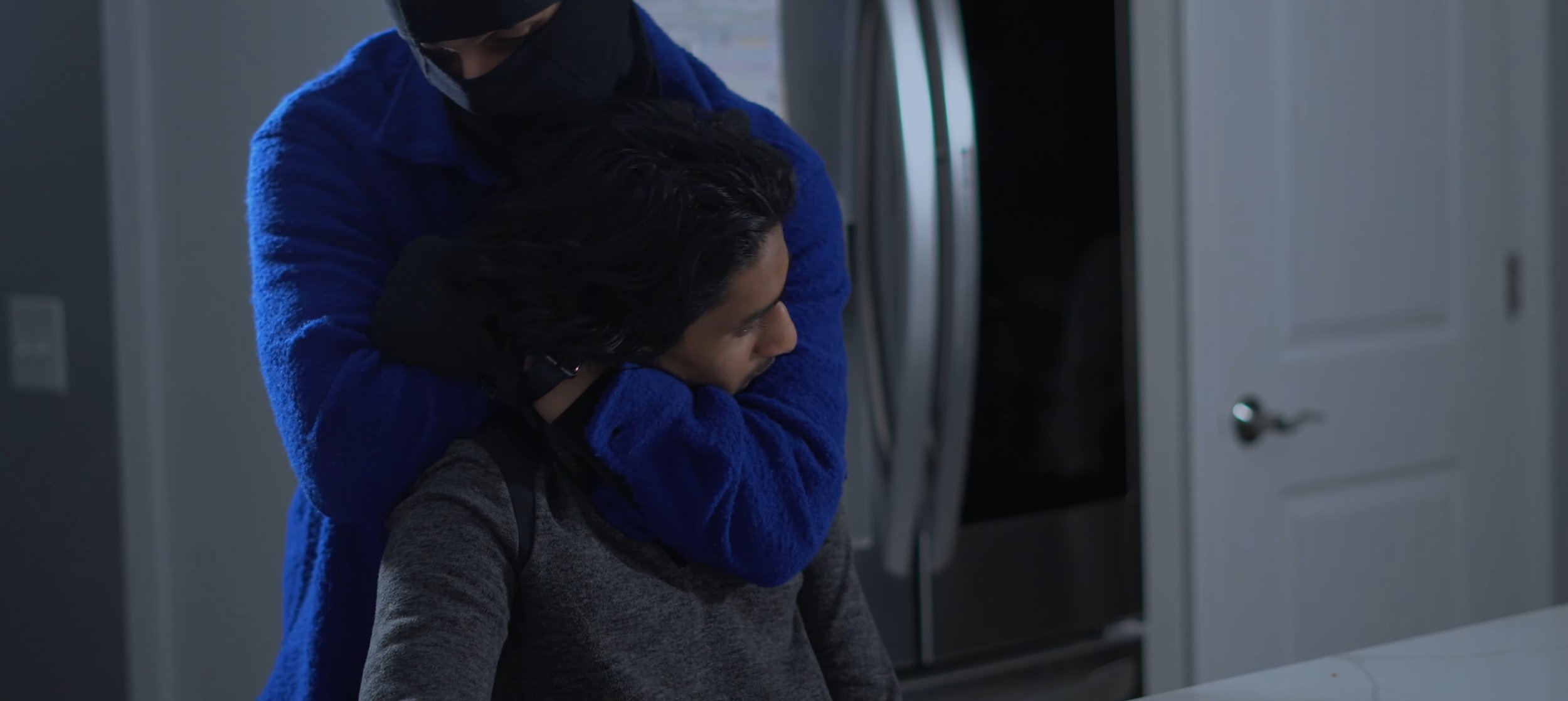 A person in a blue jacket comforting a young person with black hair, who appears upset, in a home setting near a refrigerator and a closed door.