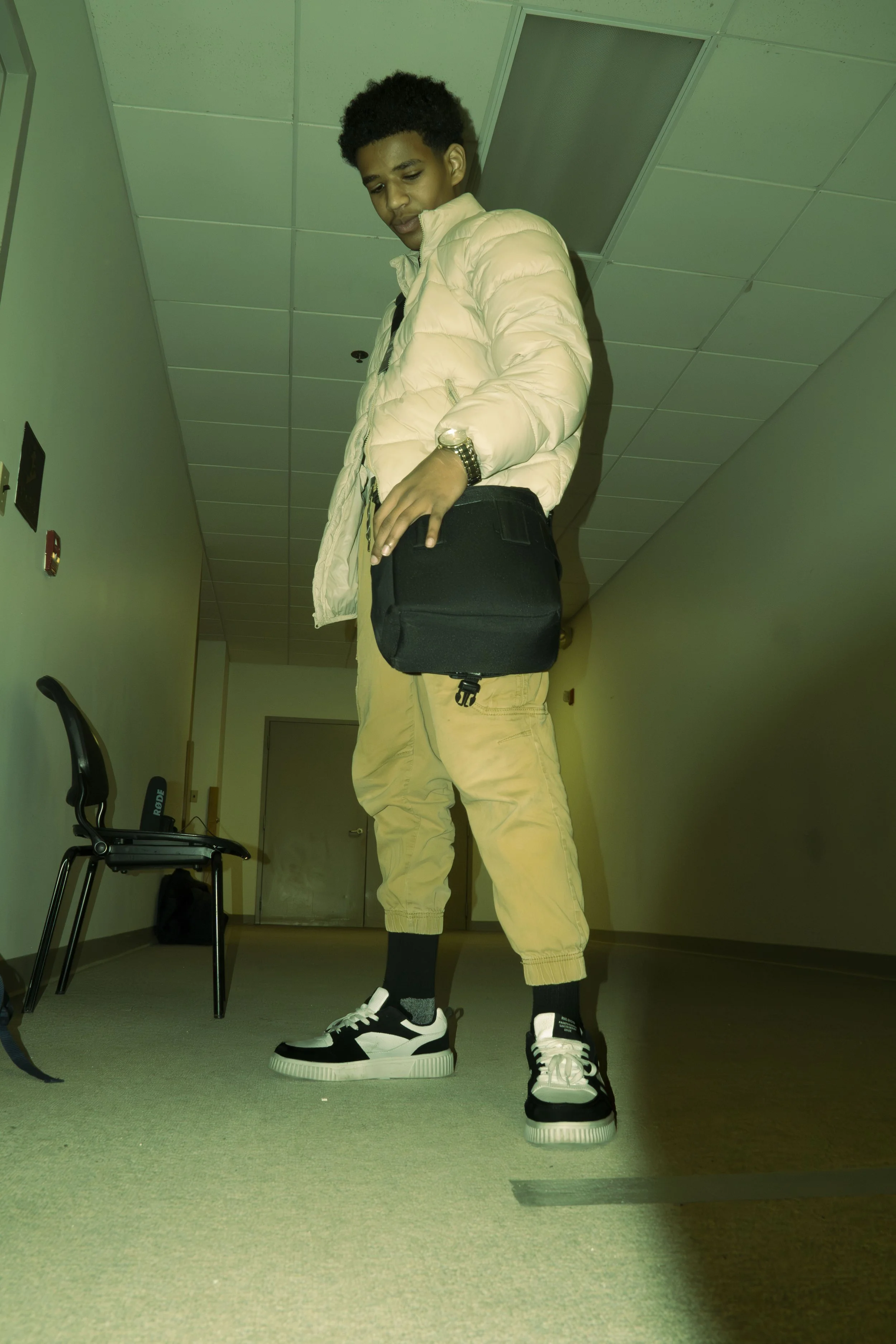A young man with dark curly hair wearing a light-colored puffer jacket, beige pants, black and white sneakers, and a black backpack, standing in a hallway.