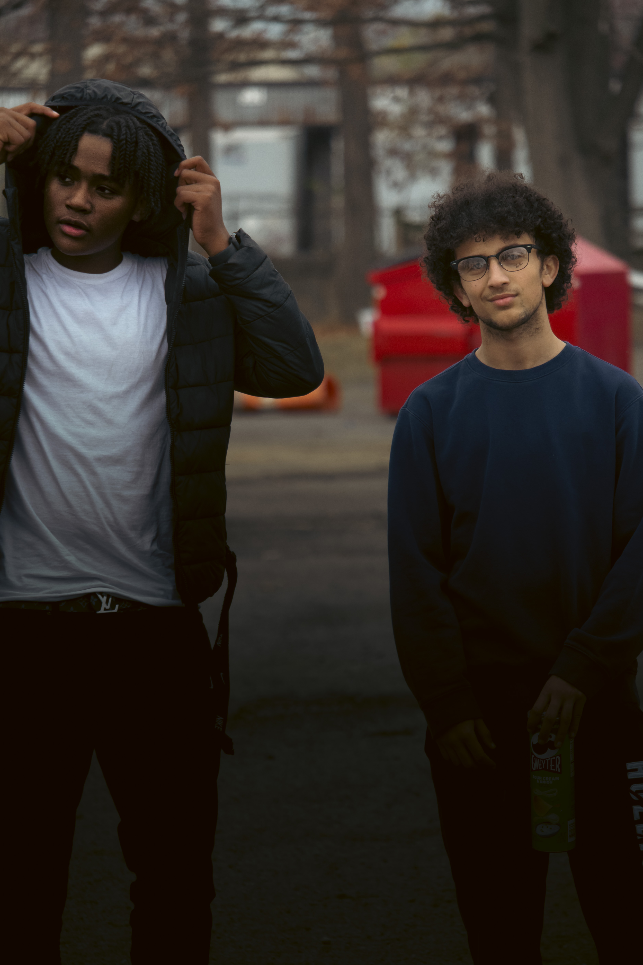 Two young men standing outdoors on a cloudy day, one adjusting his hood, the other holding a can of whipped topping, with red storage units and trees in the background.