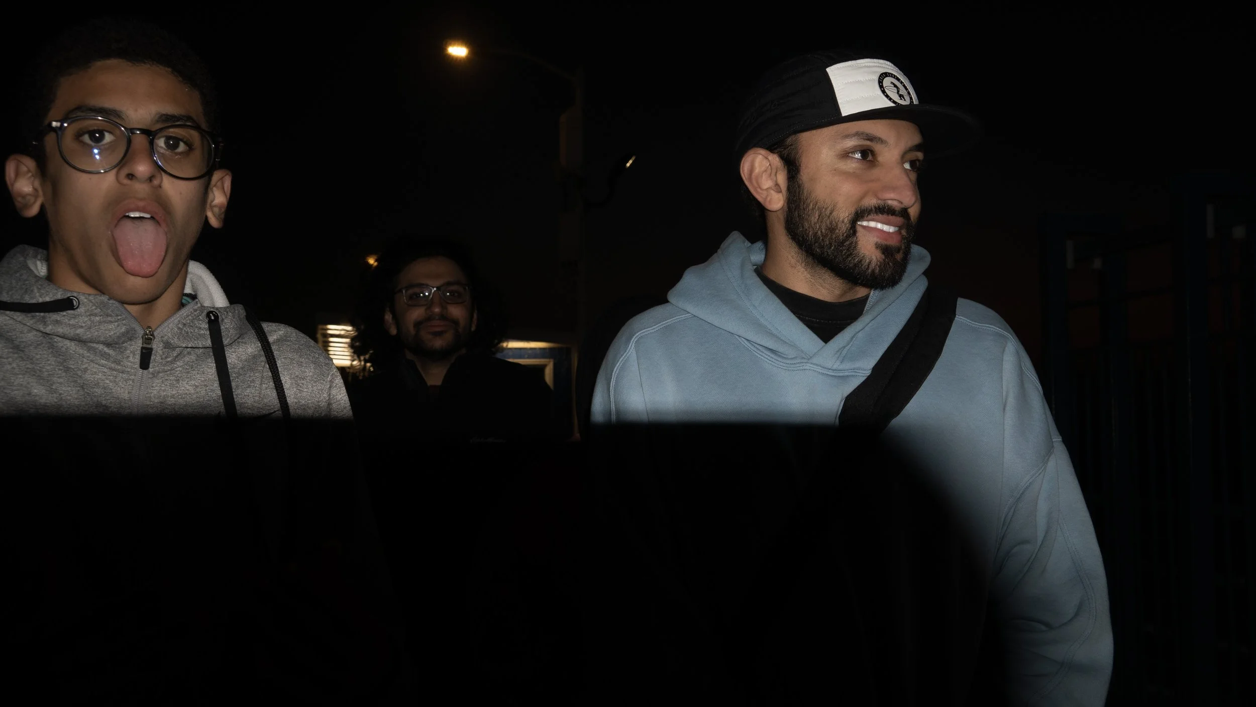 Three men walking at night, one in the foreground sticking tongue out, wearing glasses and a grey hoodie; another in the middle background with glasses and long hair, and third in the right foreground smiling, wearing a black and white cap and a blue