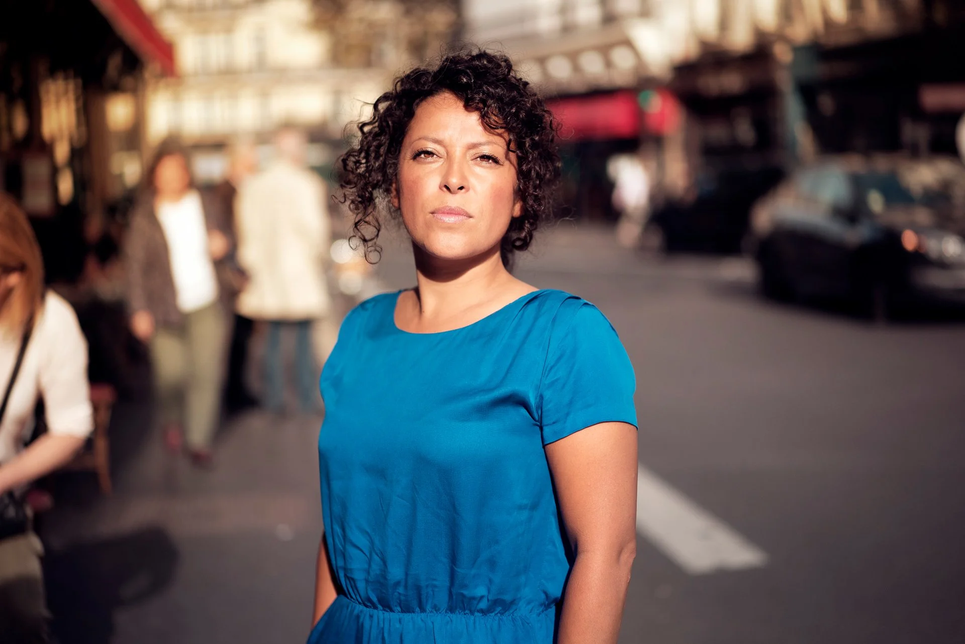 A woman with short curly dark hair and light skin is standing outdoors on a city street, wearing a blue dress, looking directly at the camera with a serious expression. Blurred pedestrians and vehicles are visible in the background.