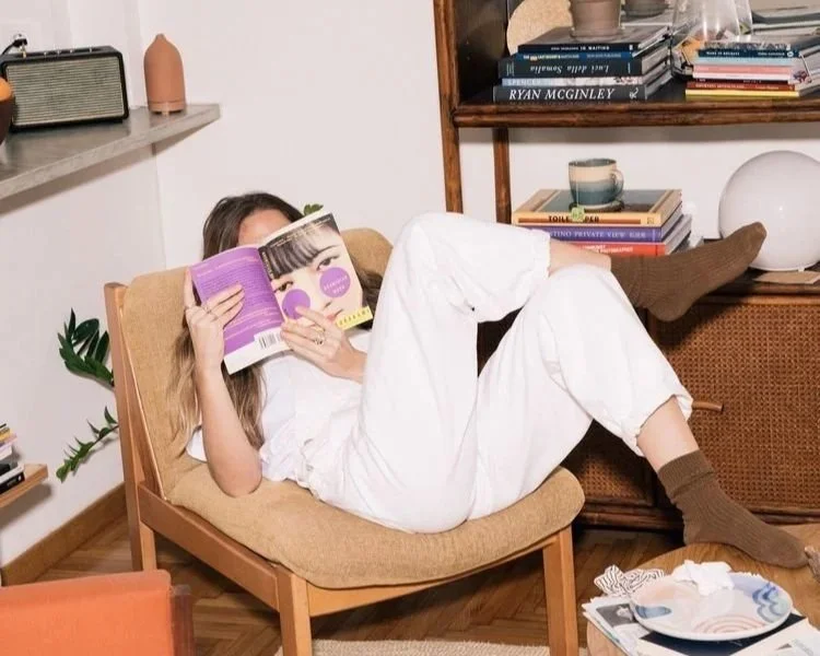 A woman with long hair, dressed in white, is lying on a tan armchair, reading a magazine, with her legs resting on a sideboard. She is wearing brown socks. The room has books on shelves, a potted plant, and various decorative items.