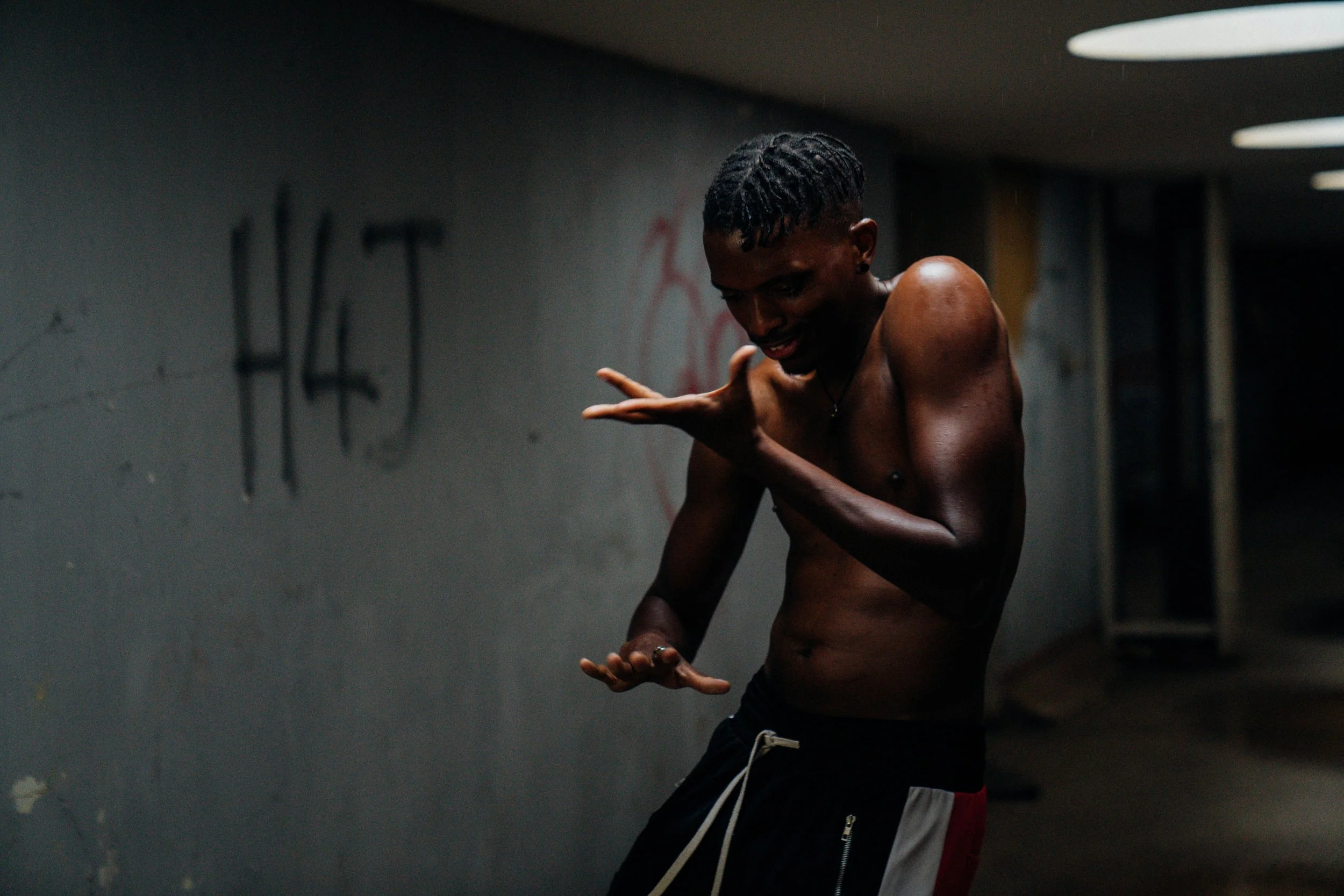 A young man stands in a dimly lit, graffiti-covered room, suggesting physical presence, energy, and human agency.
