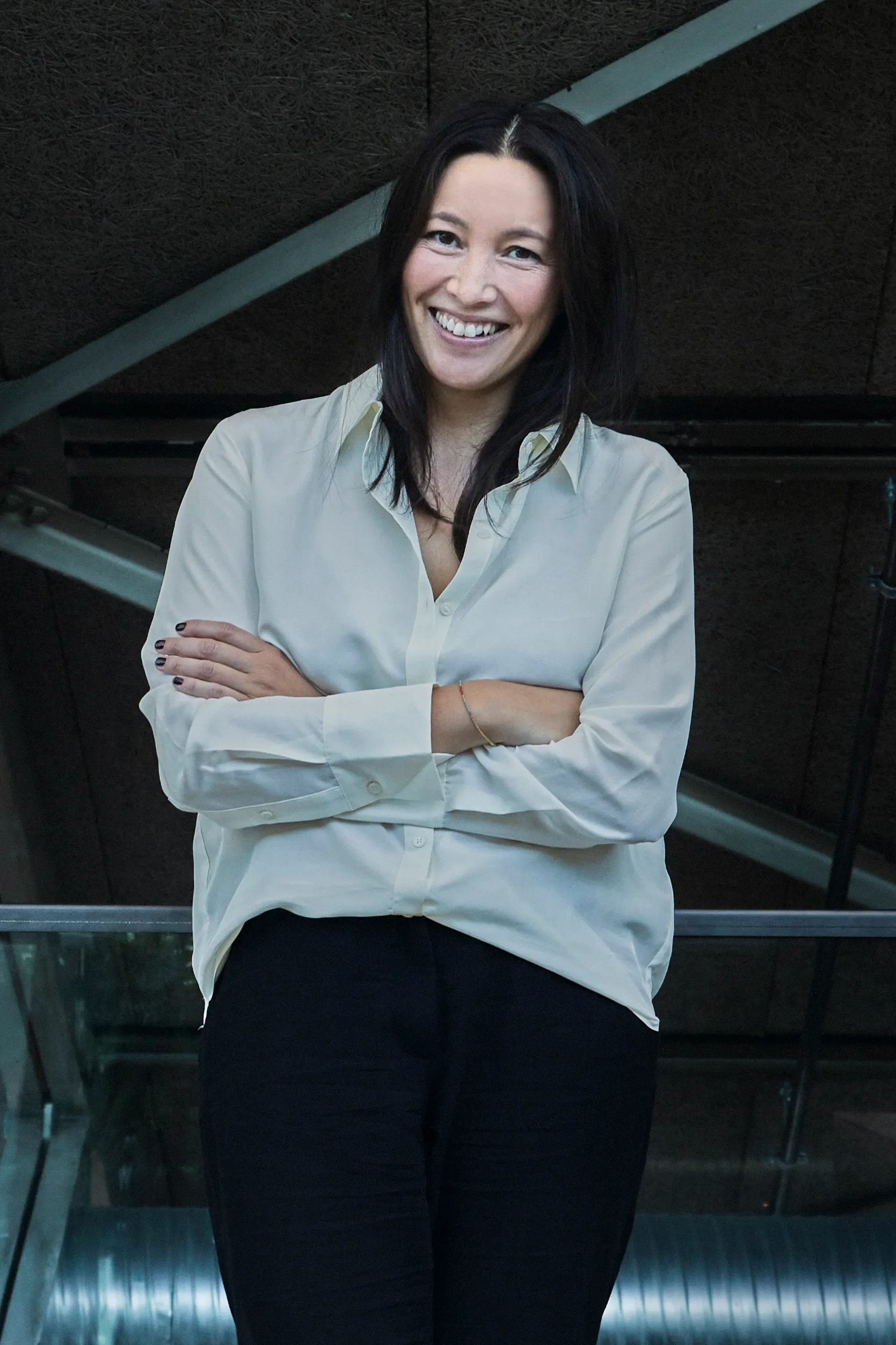 A woman with long black hair smiling, wearing a off-white button-up shirt and black pants, standing with arms crossed in front of a dark ceiling with metal beams.