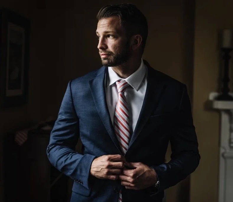 Man in a blue suit adjusting his jacket, wearing a white shirt and striped red tie, in a dimly lit room.