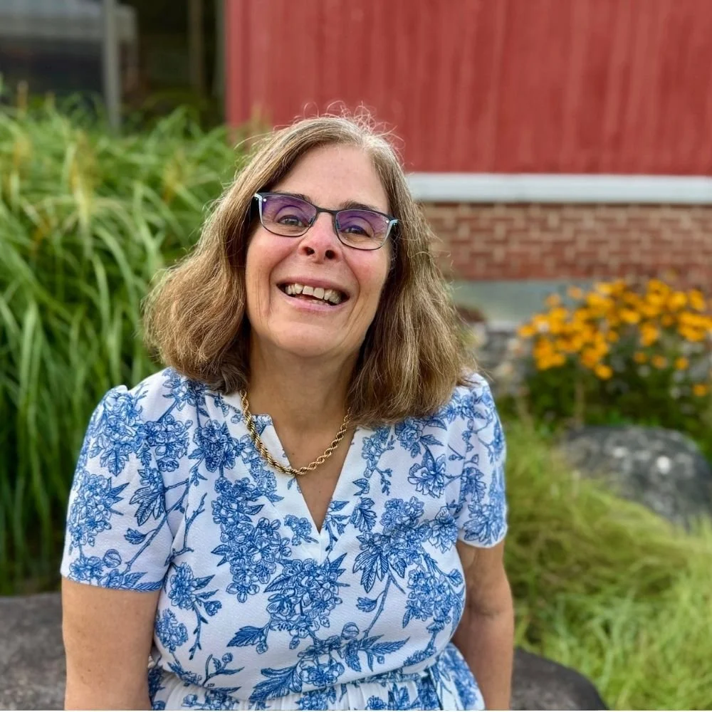 A woman with brown hair, Rep. Zoe Manos, smiling at the camera with yellow flowers and green shrubbery in the background