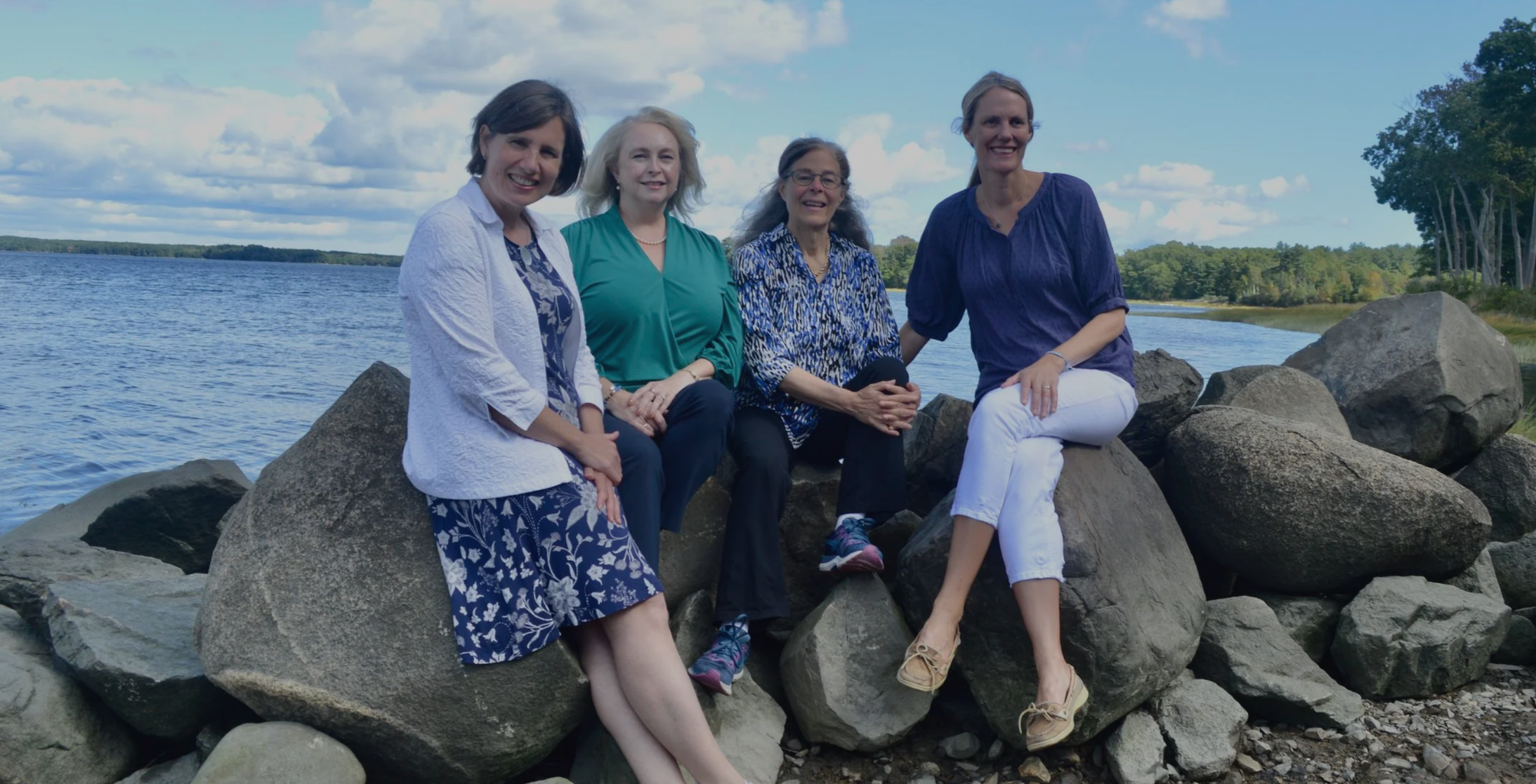 Four women sitting on rocks by a lake with trees in the background, smiling and enjoying the outdoor scenery on a partly cloudy day.