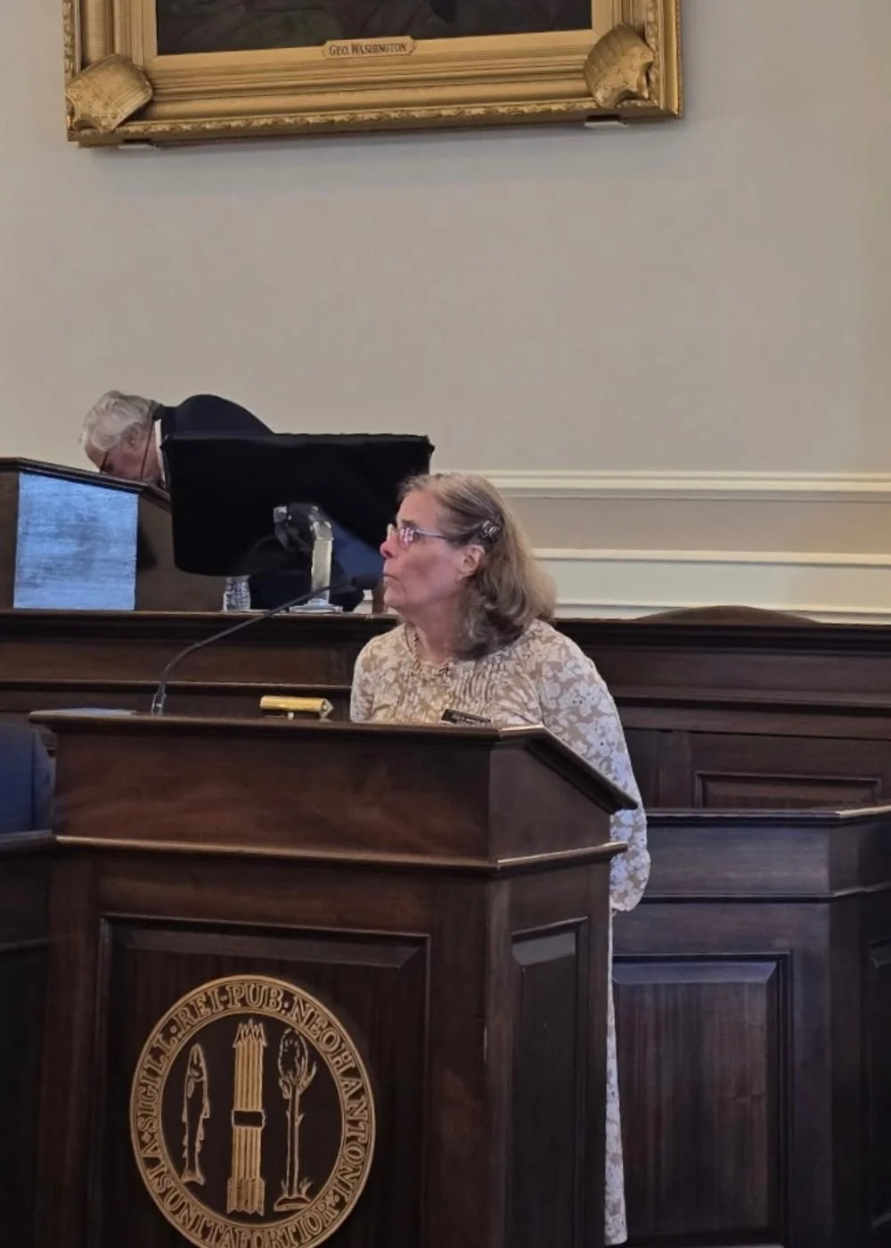 A woman with glasses and shoulder-length hair (Rep. Zoe Manos) speaking at a wooden podium with a seal, in a formal setting with a man in the background and a large framed portrait hanging on the wall.