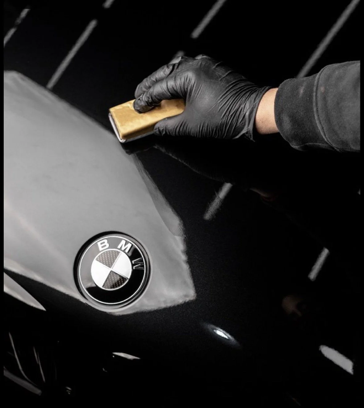 Person wearing black gloves polishing the hood of a BMW car with a cloth in a garage or workshop.