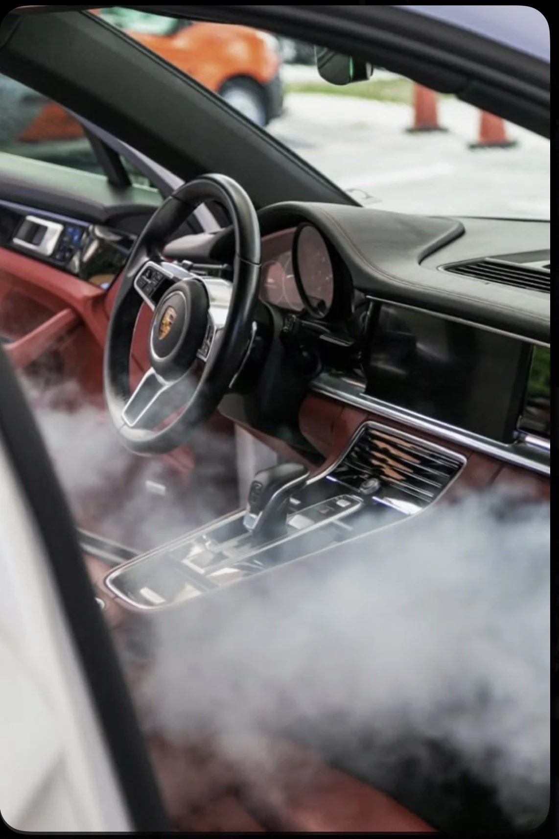 The interior of a Porsche vehicle, showing a steering wheel with the Porsche emblem, an automatic gear shift, and a dashboard with a digital display, visible through a car window.