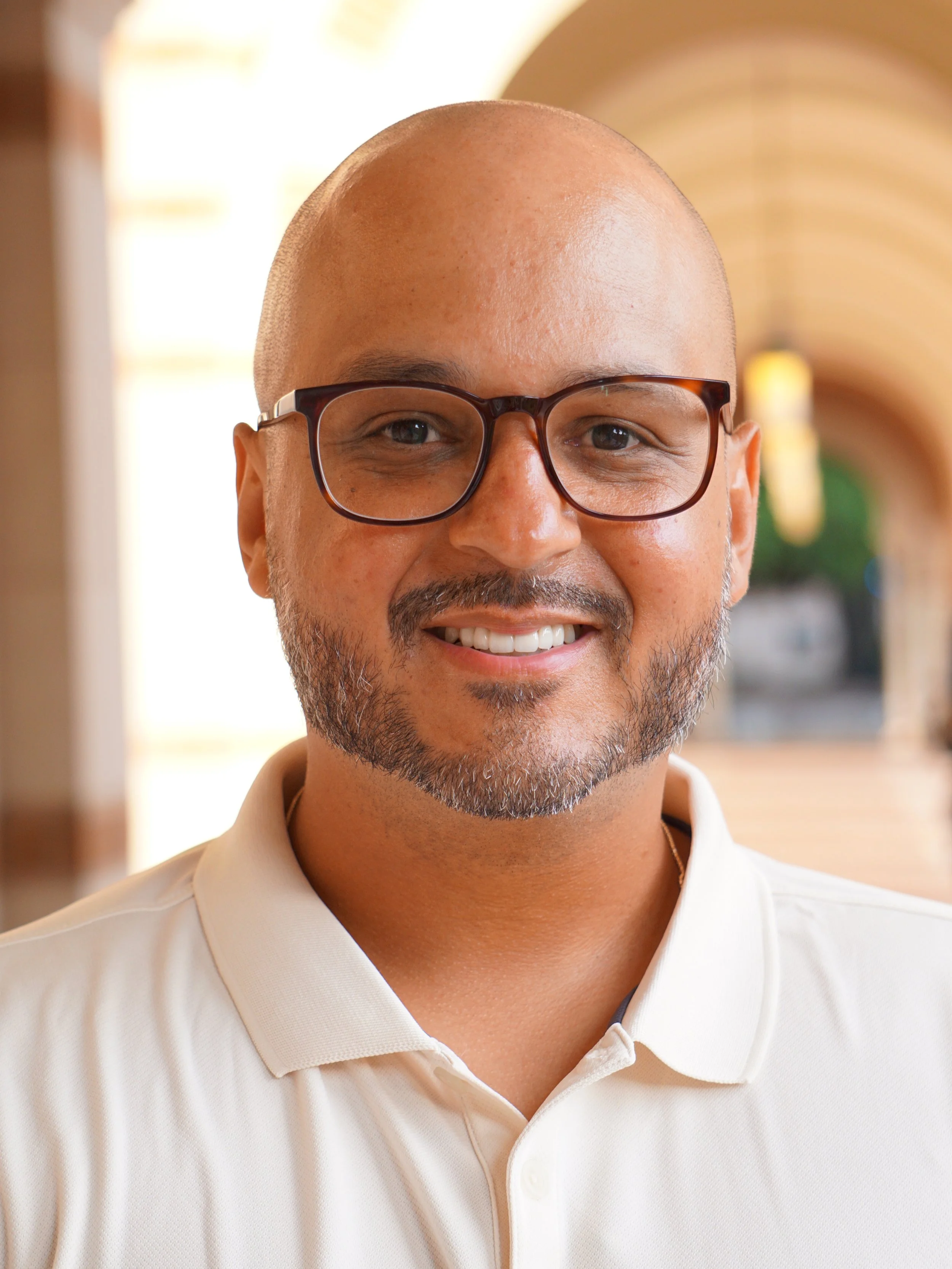 A smiling man wearing glasses and a light-colored polo shirt, standing in a bright indoor space with arches and greenery.